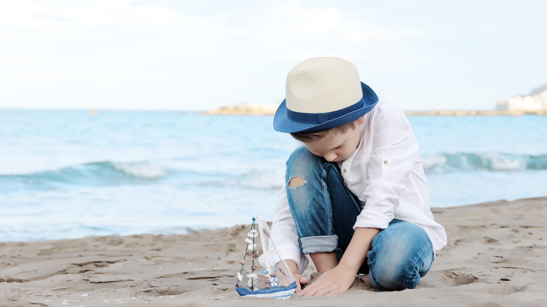 Маленький мальчик в шляпе играет с игрушечным корабликом на песке