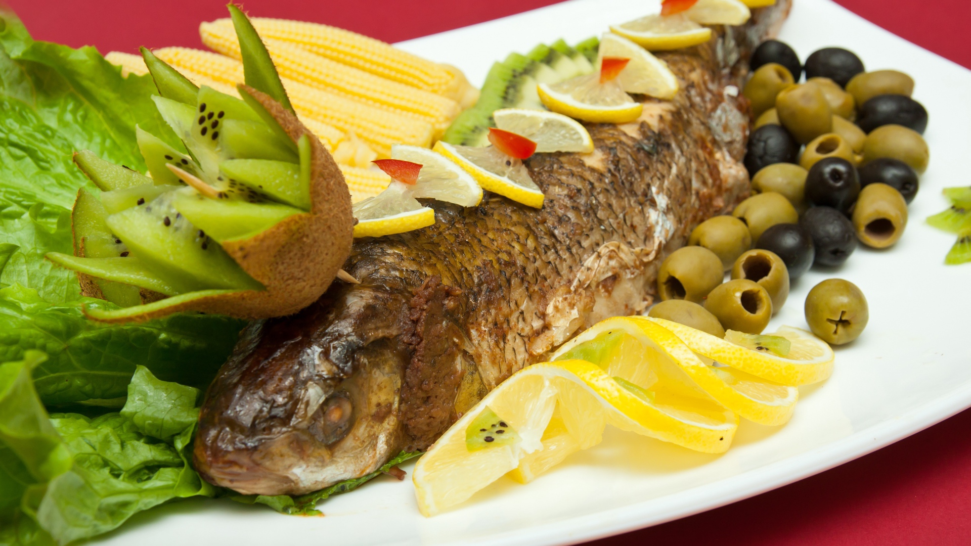 Запеченная рыба с оливками, лимоном, киви, кукурузой и листьями салата на белой тарелке