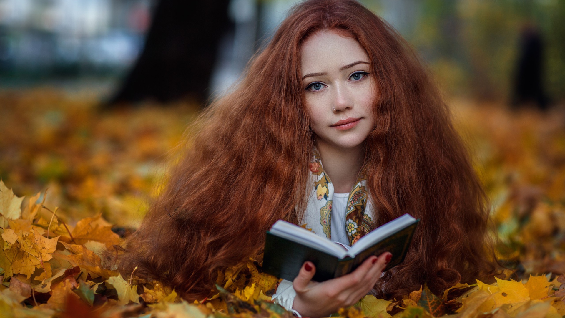 Рыжеволосая девушка с книгой лежит на опавших листьях осенью 