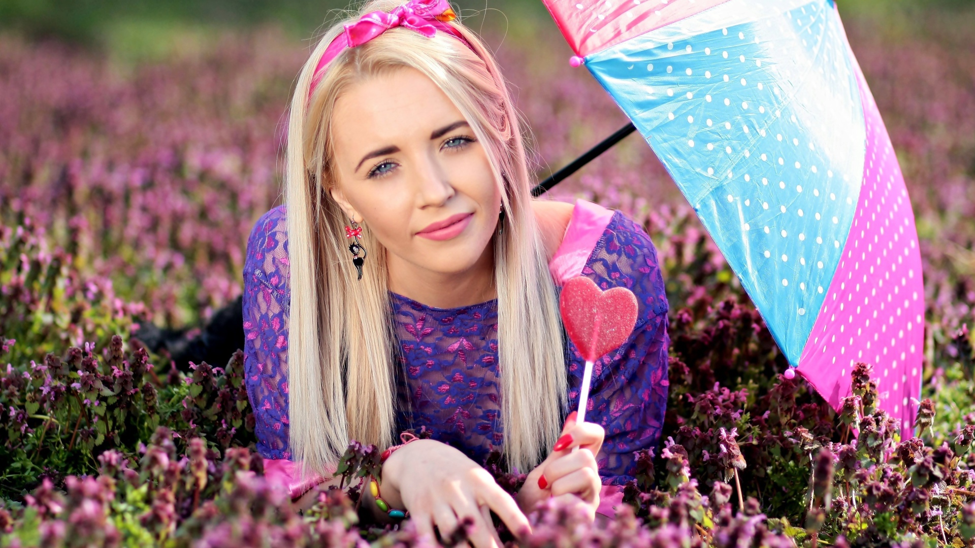 Молодая девушка блондинка лежит на поляне с сиреневыми цветами под разноцветным зонтом