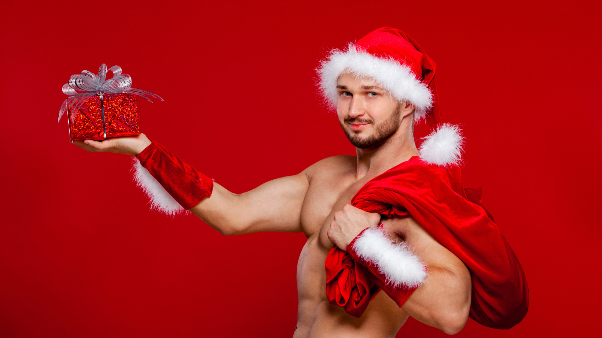 Мужчина в новогоднем костюме Санта Клауса с мешком и подарком в руке на красном фоне