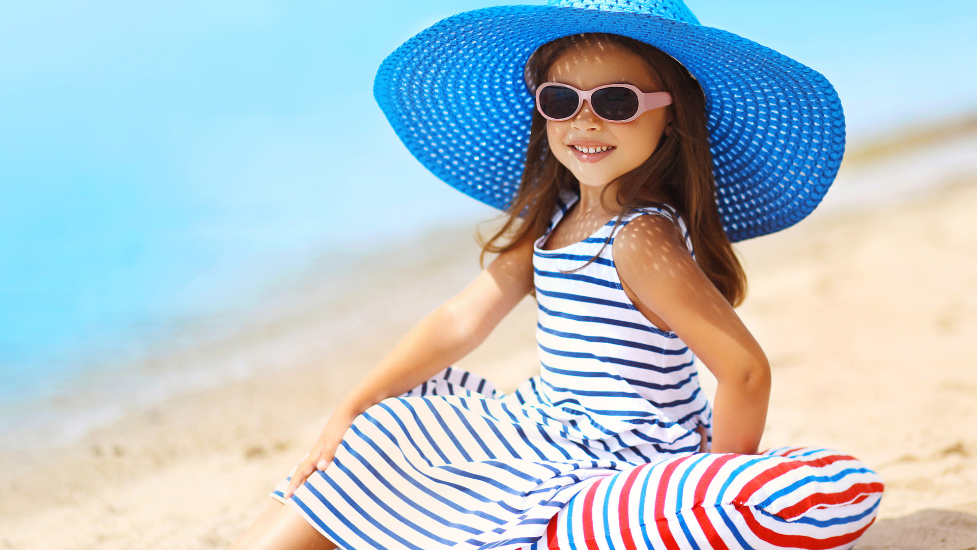 Маленькая девочка сидит на подушке на пляже в большой синей шляпе