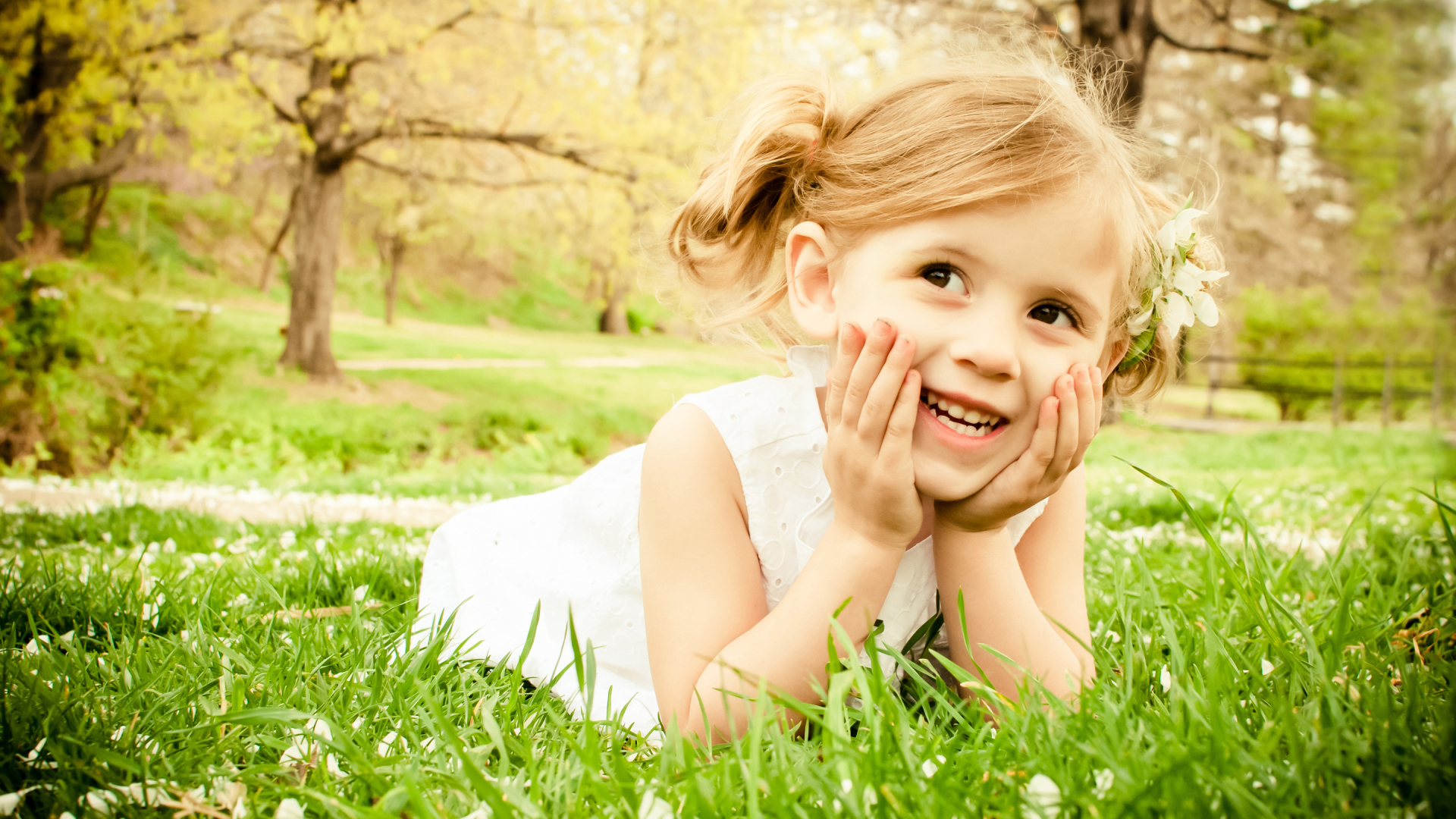 Улыбающаяся девочка в белом платье лежит на зеленой траве