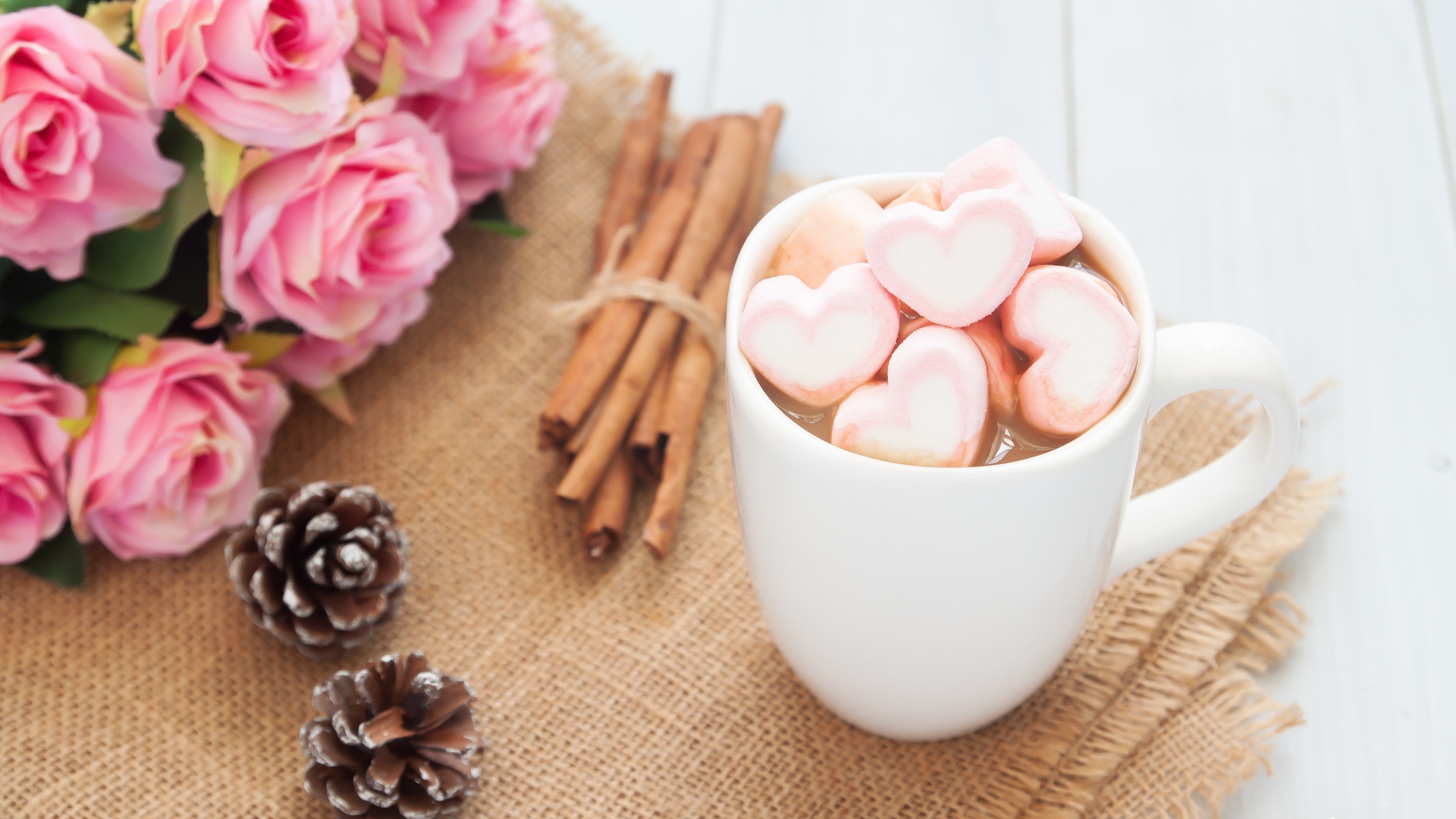 Чашка какао с маршмеллоу на столе с корицей, шишками и букетом розовых роз