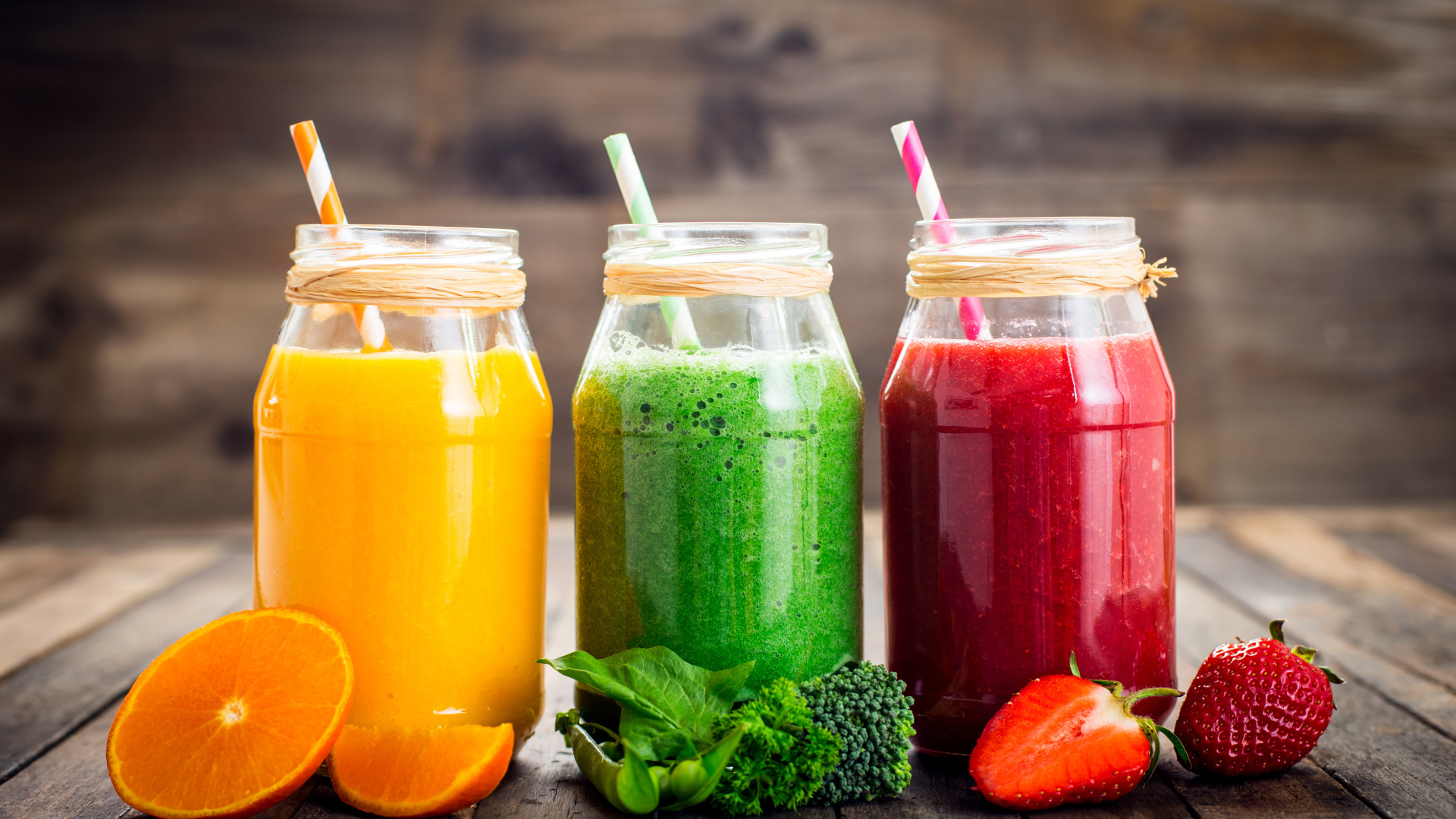 Три банки смузи с трубочкой на столе с апельсином, брокколи и клубникой