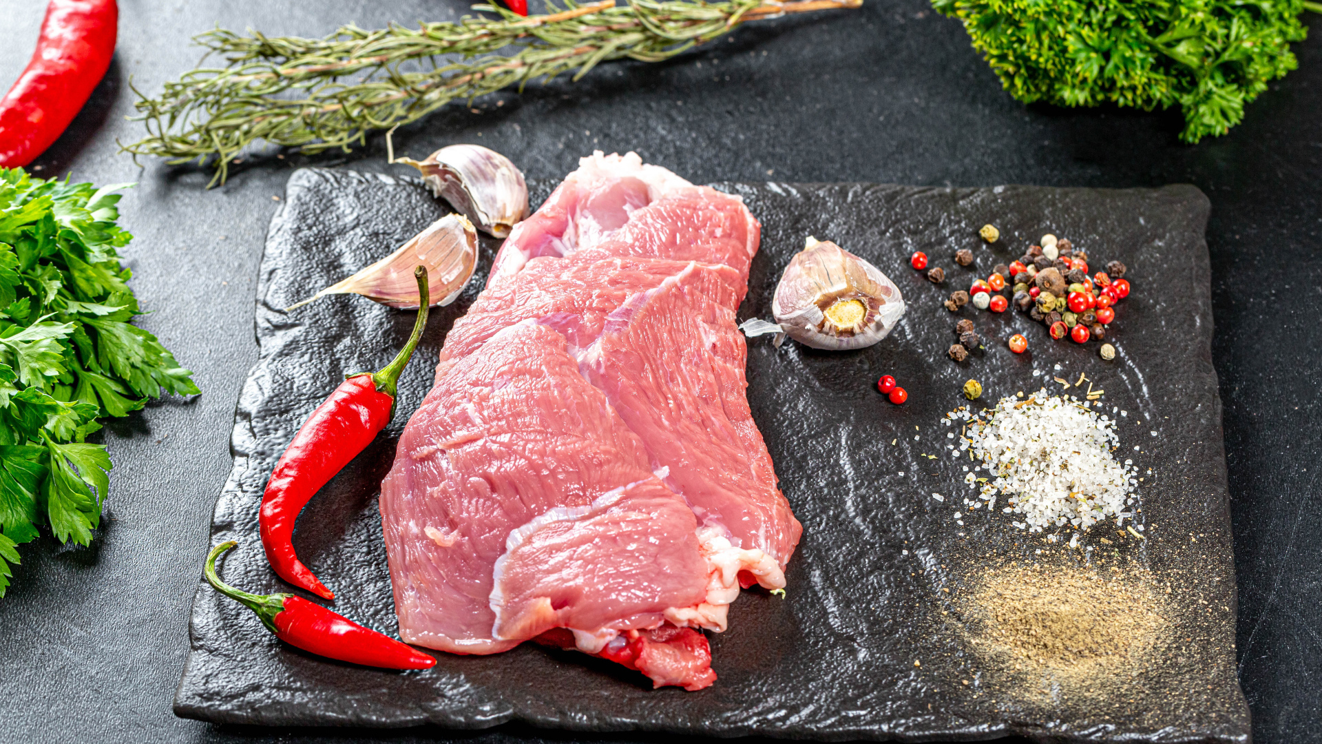 Кусок мяса на столе с острым перцем, чесноком и зеленью
