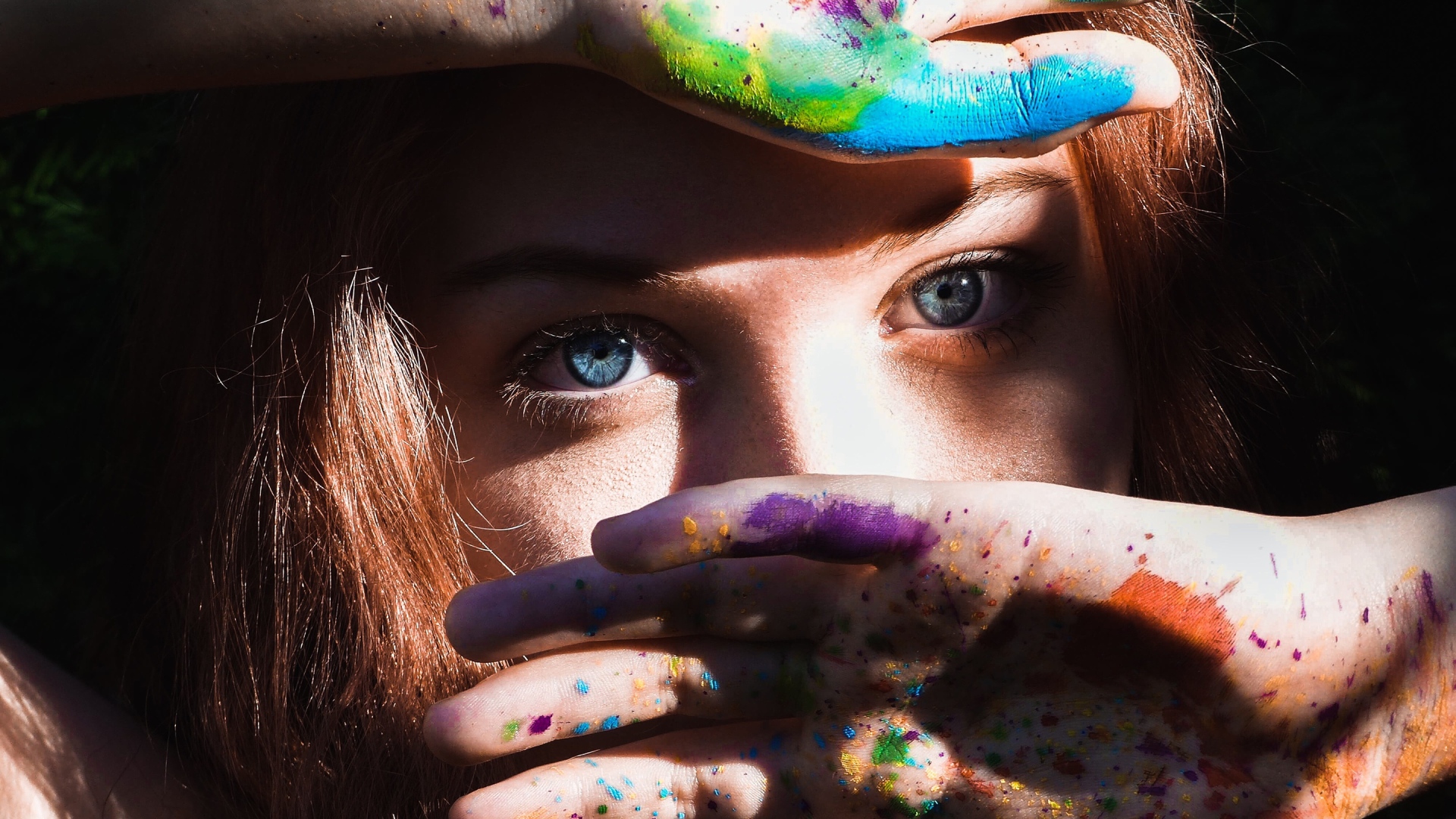 Красивая голубоглазая девушка с руками в разноцветной краске у лица