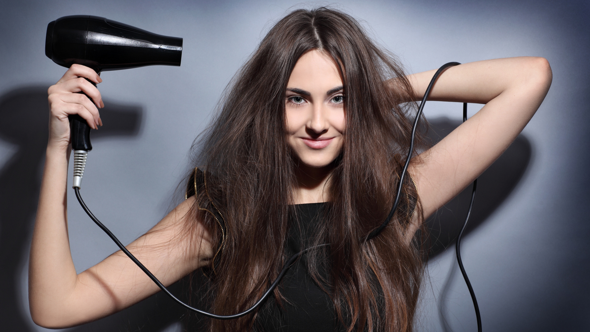 Длинноволосая девушка сушит волосы феном на сером фоне