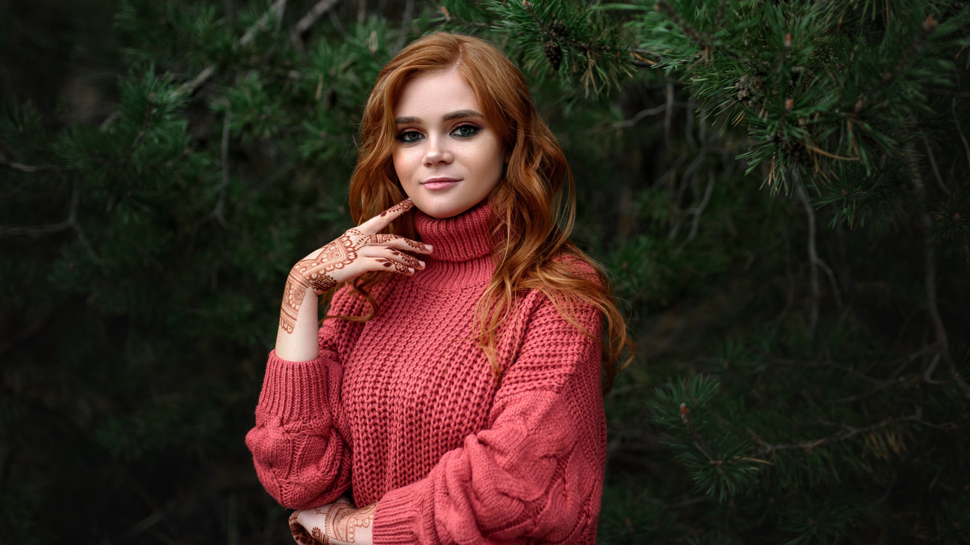 Рыжеволосая девушка в розовом свитере стоит у ели