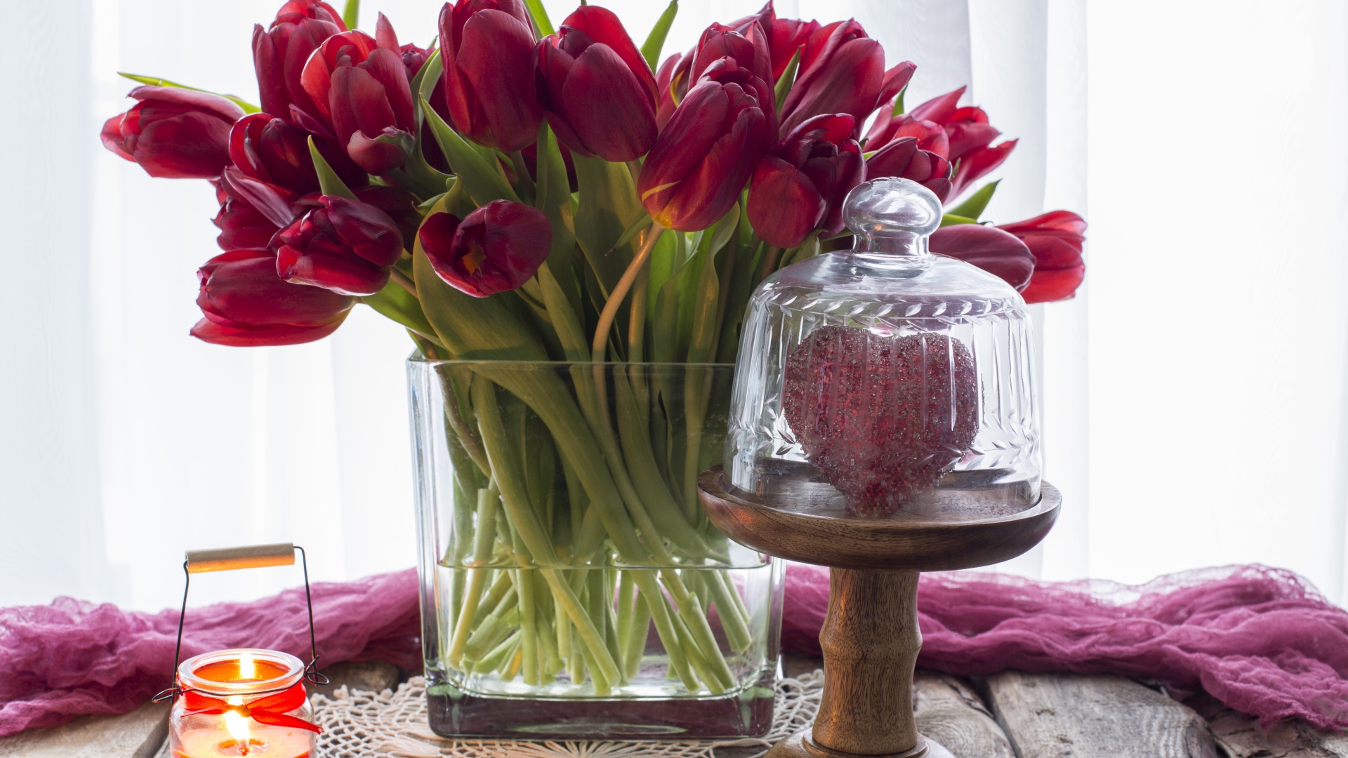 Букет красных тюльпанов в стеклянной вазе на столе со свечой и сердцем 