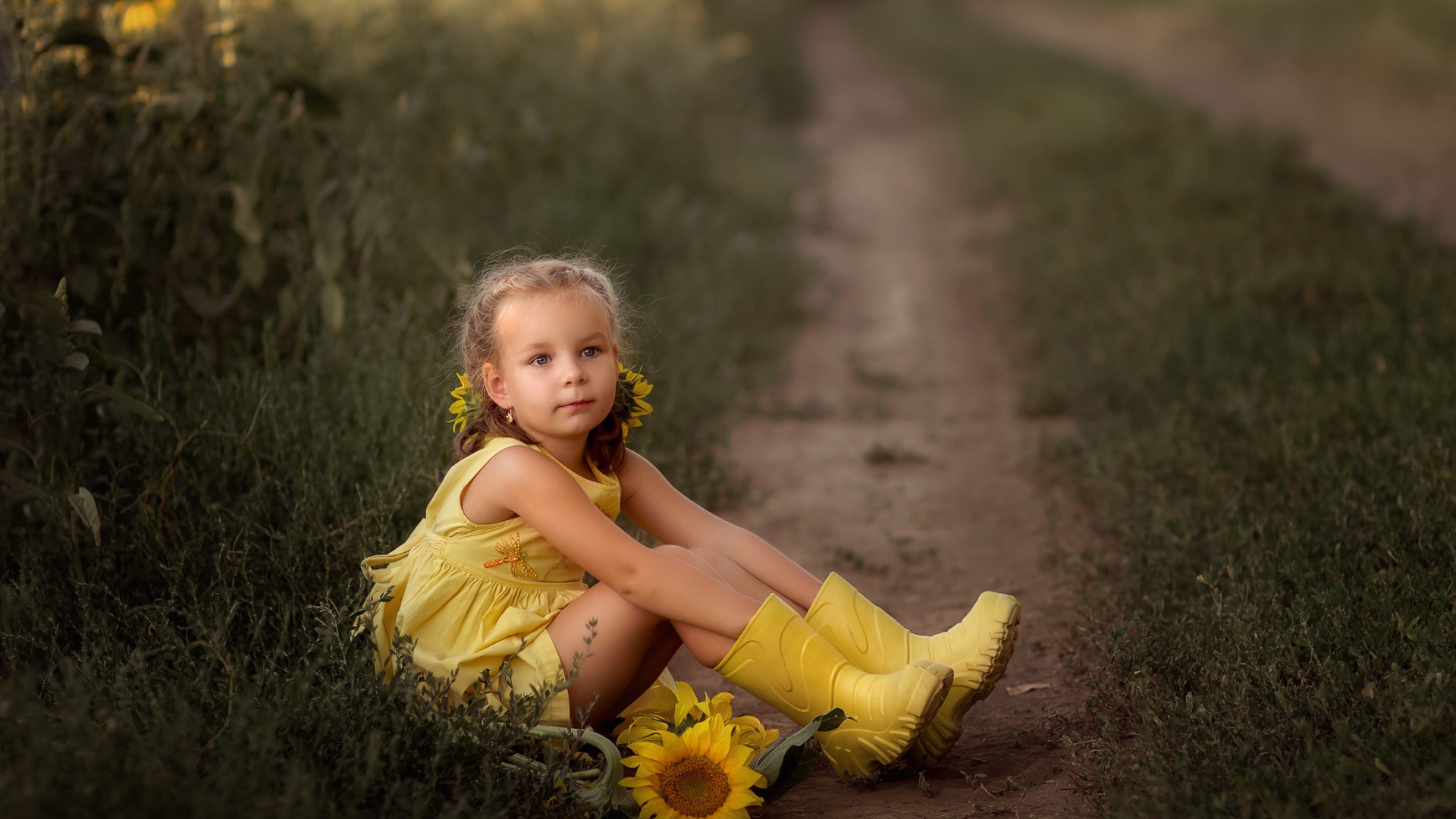 Маленькая девочка в желтом платье сидит на земле с подсолнухами
