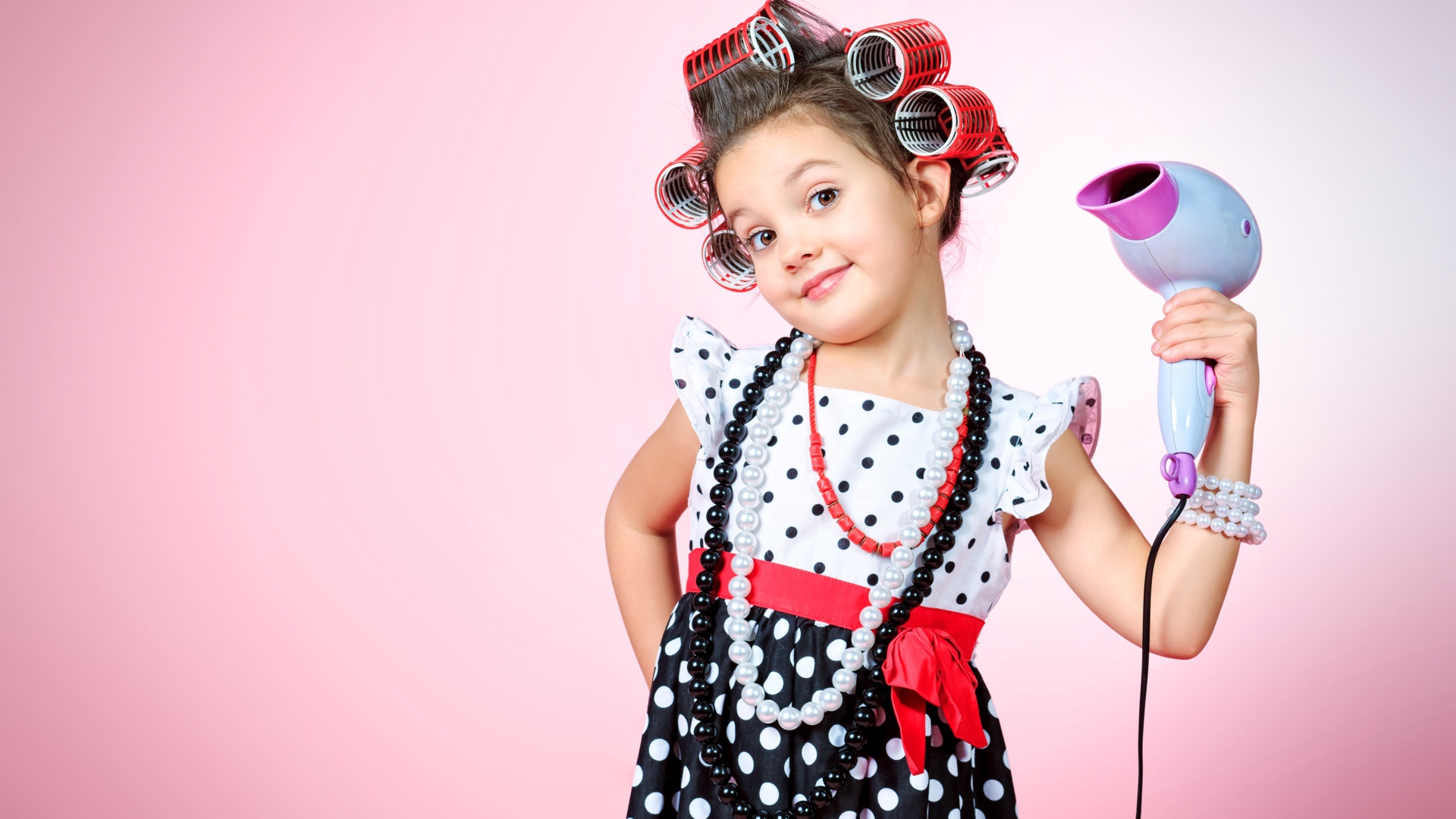 Маленькая девочка с бигудями на голове с феном в руке на розовом фоне