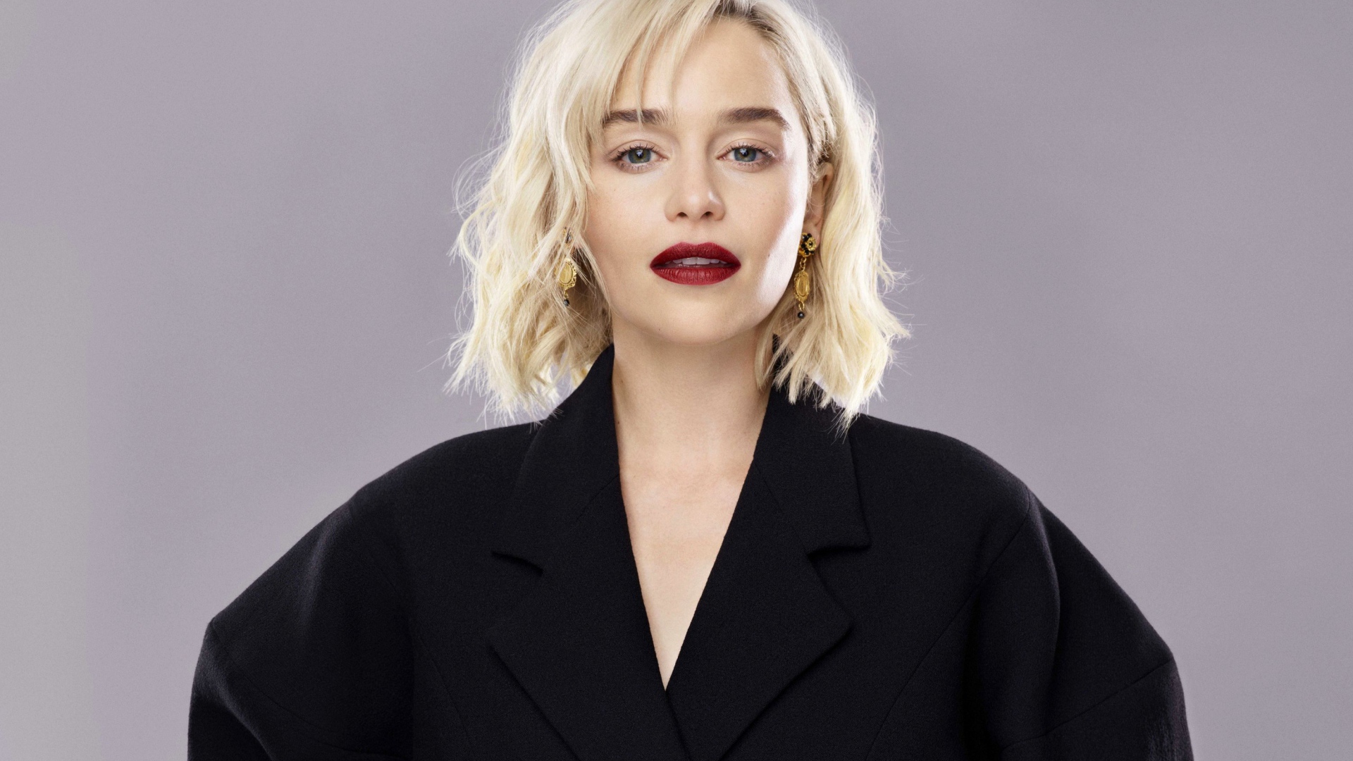 Яркая блондинка, актриса Эмилия Кларк в черном пальто на сером фоне