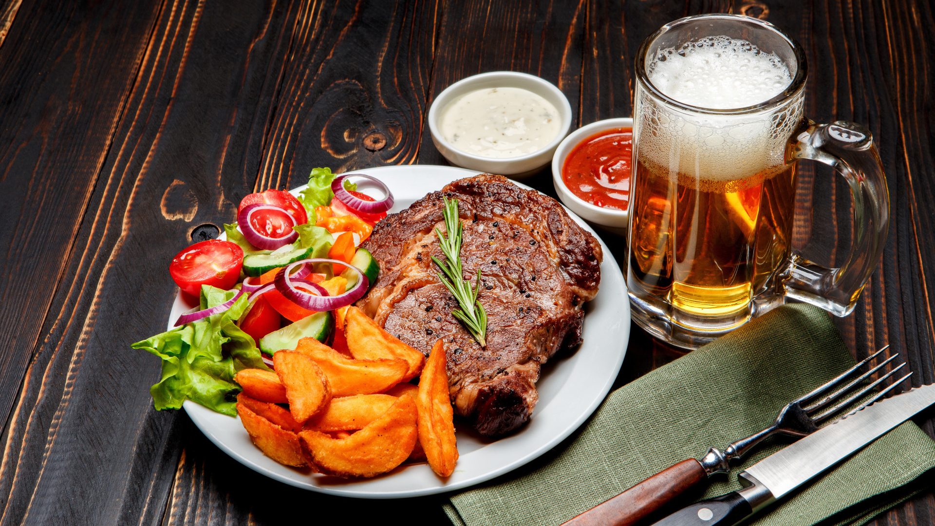 Кусок мяса на тарелке с картофелем и салатом на столе с соусом и пивом