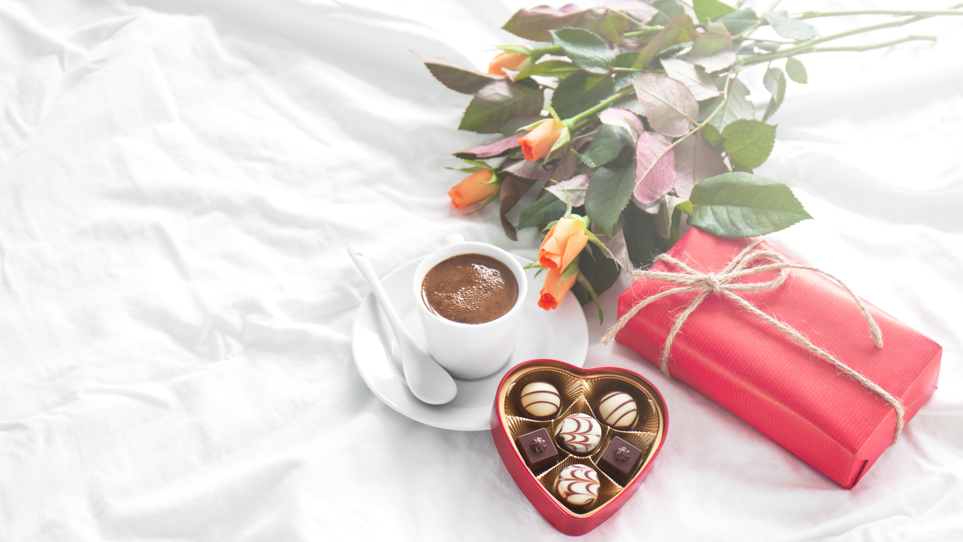 Чашка кофе на белой кровати с конфетами, подарком и букетом роз