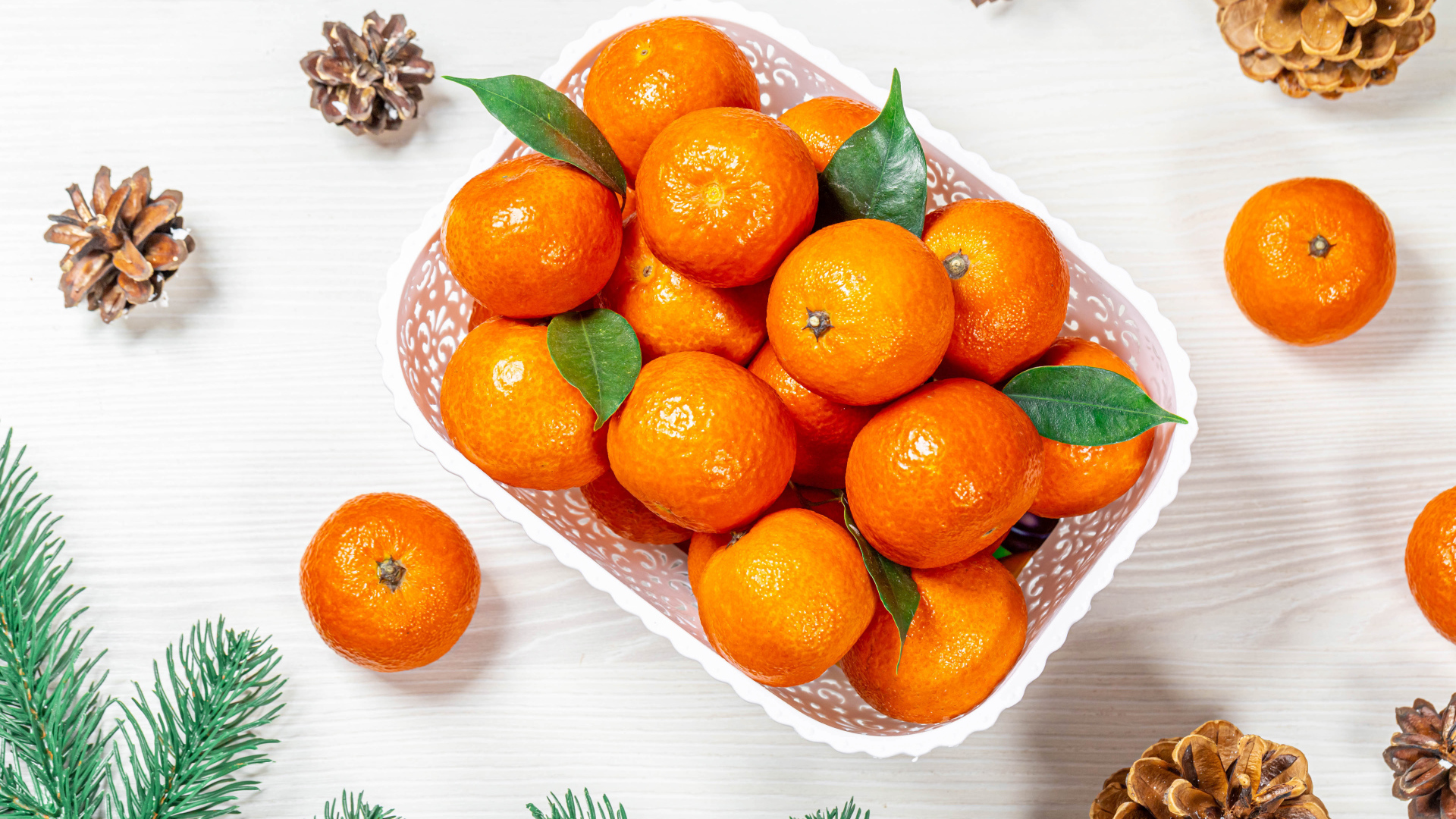 Много спелых мандаринов в белой корзинке на столе с шишками