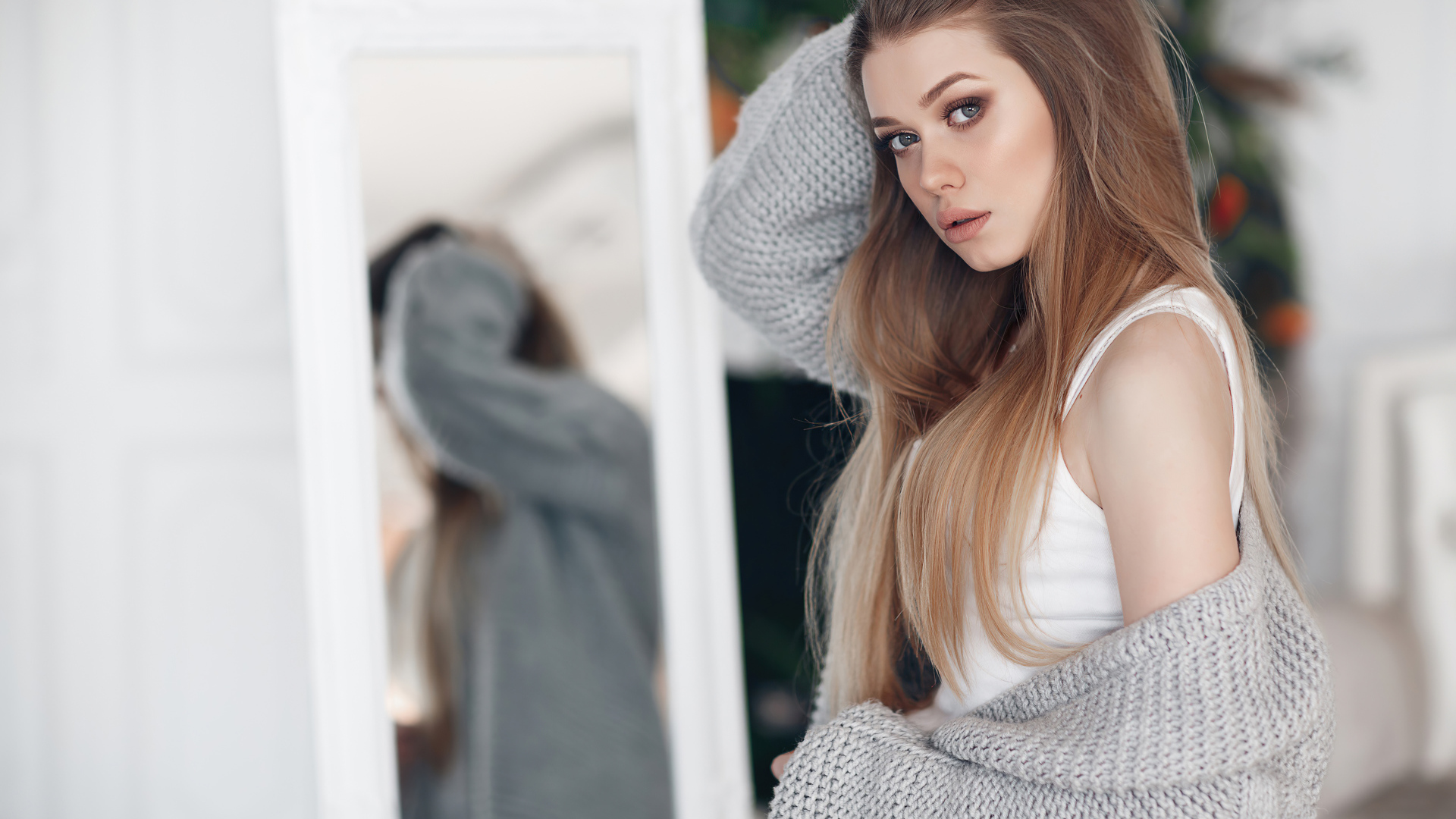 Молодая девушка в серой кофте стоит у зеркала 