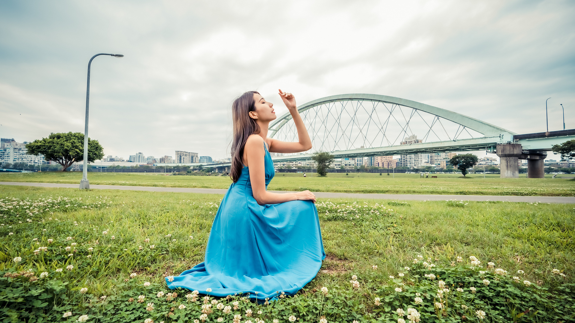 Красивая девушка азиатка в голубом платье  сидит на траве на фоне моста