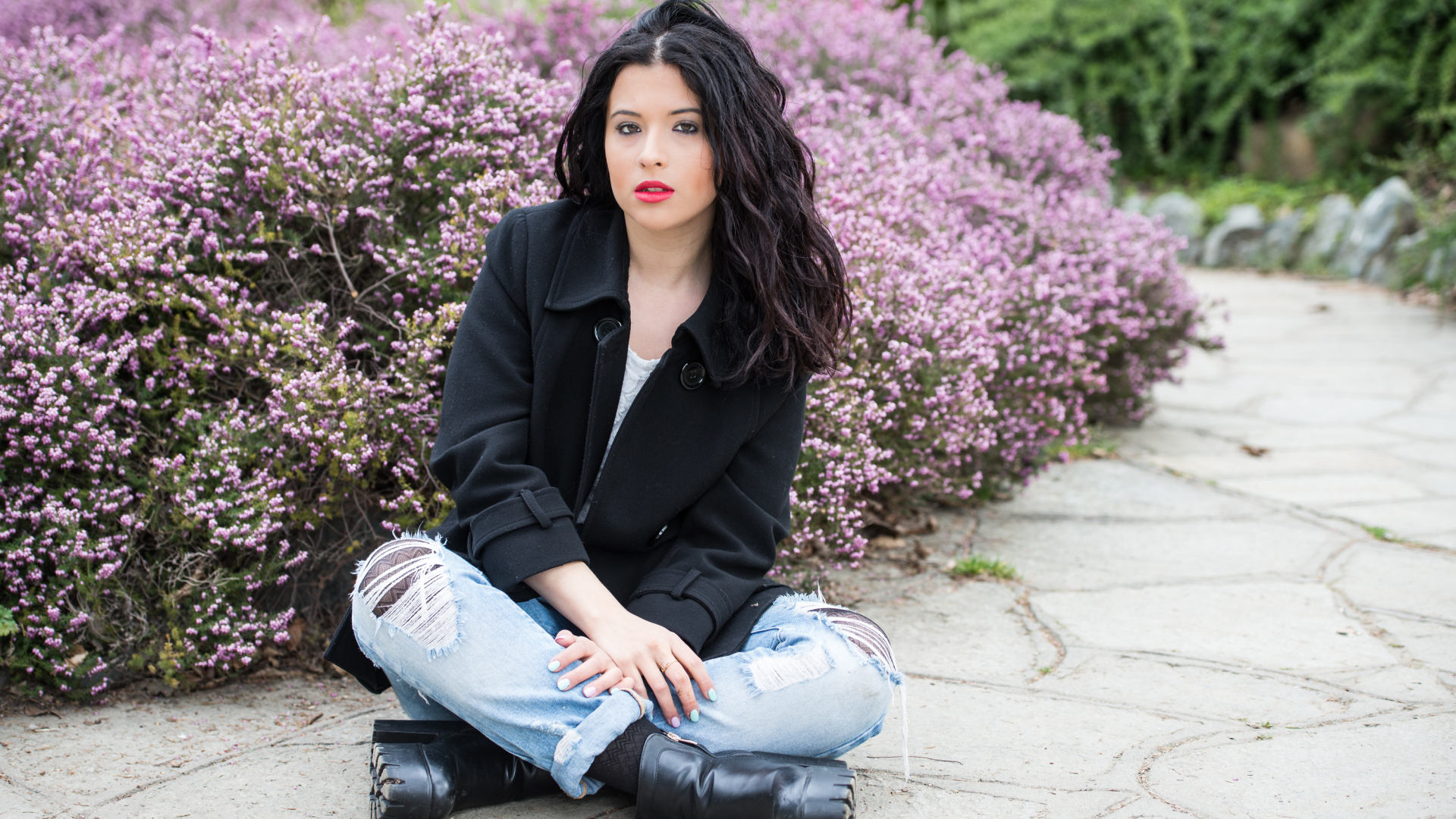 Красивая девушка в черном пальто сидит на земле у кустов