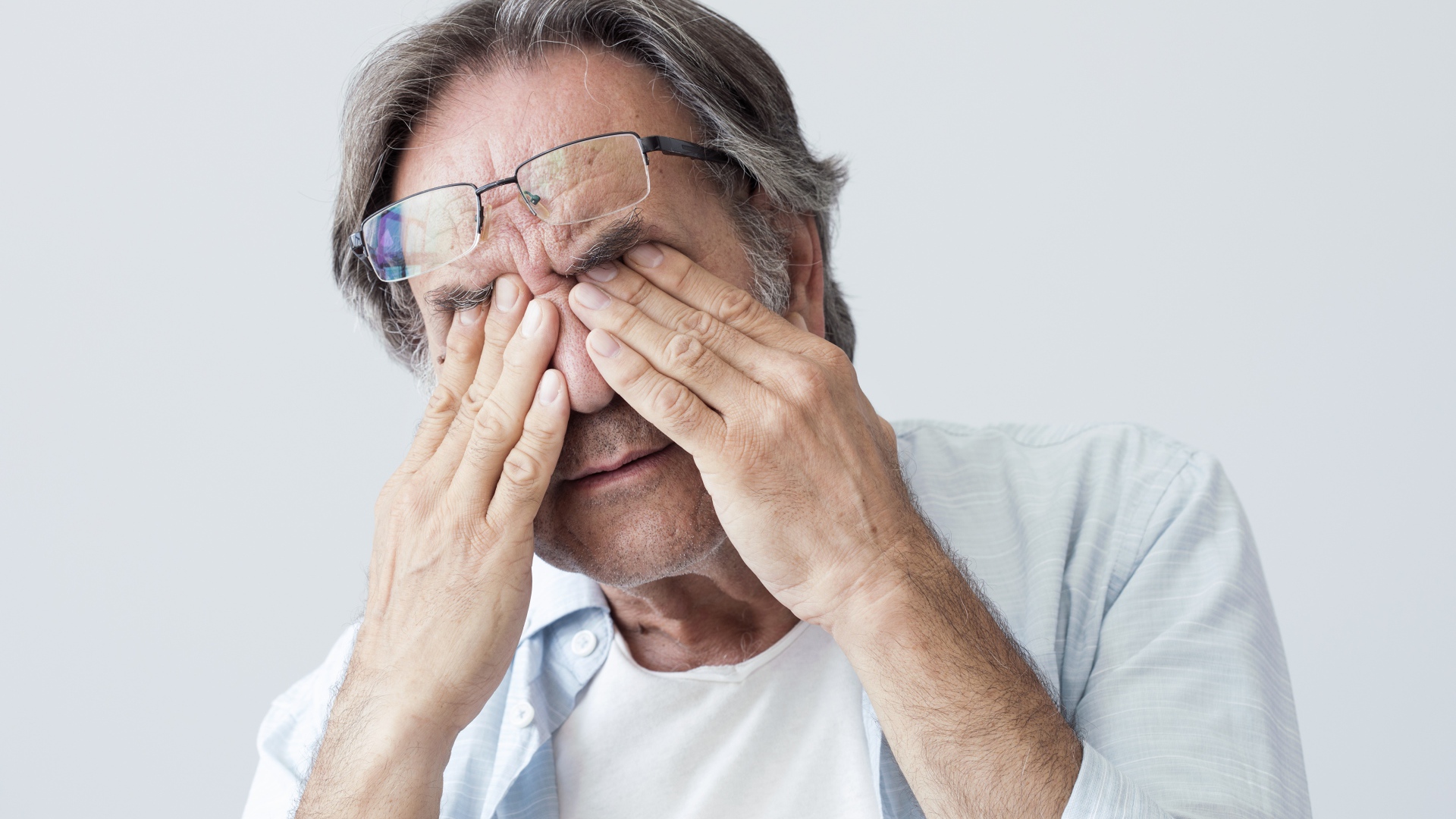 Elderly man rubs his eyes on a gray background