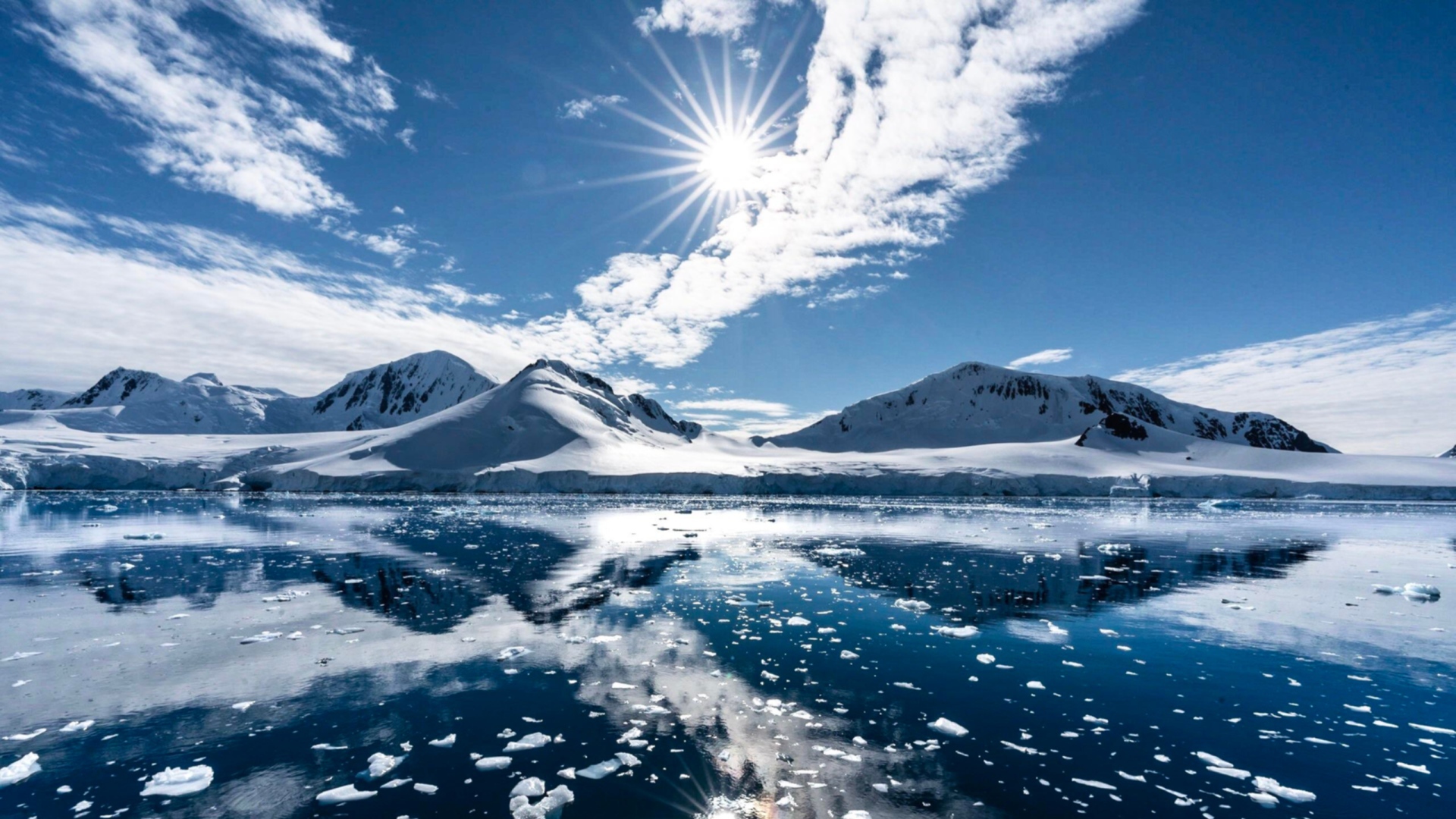 Белые облака в солнечном небе отражаются в озере у заснеженных гор 