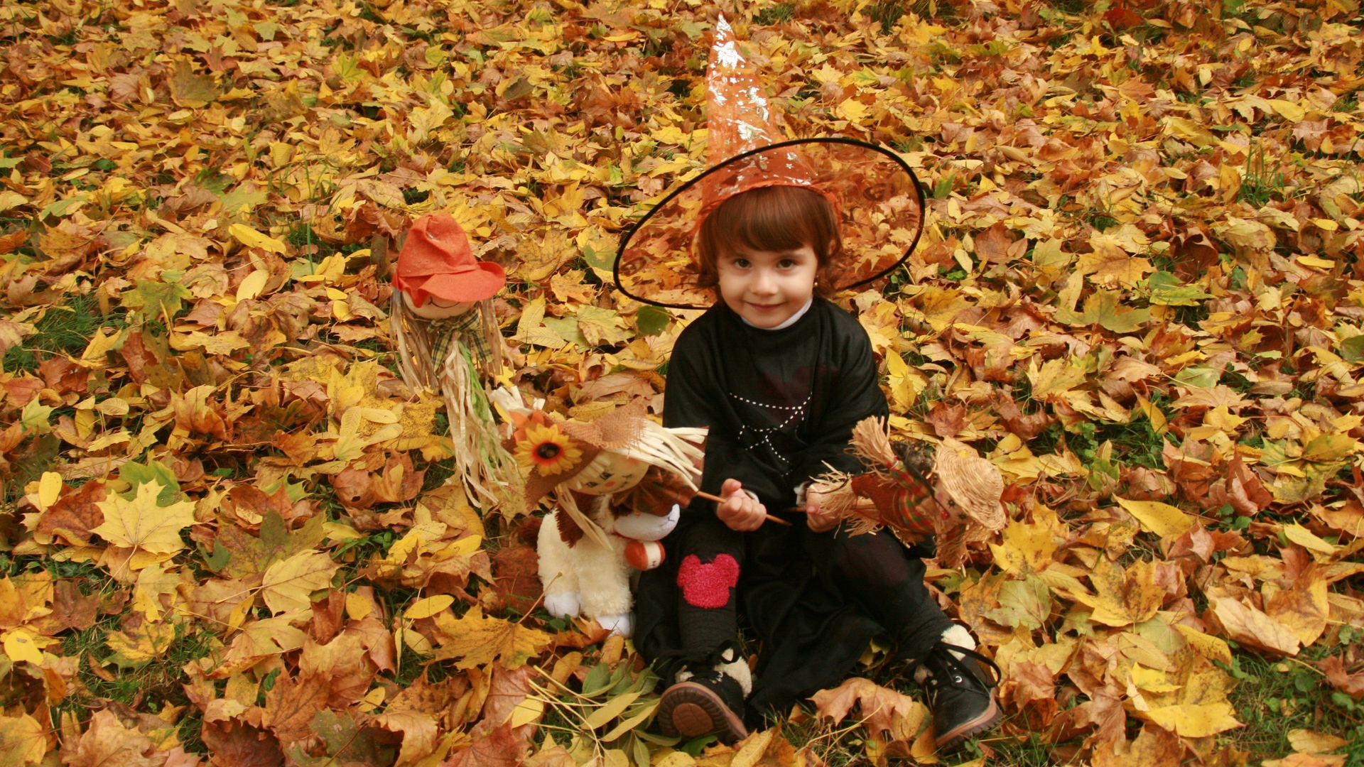 Маленькая девочка в костюме ведьмы сидит на опавшей листве на Хэллоуин