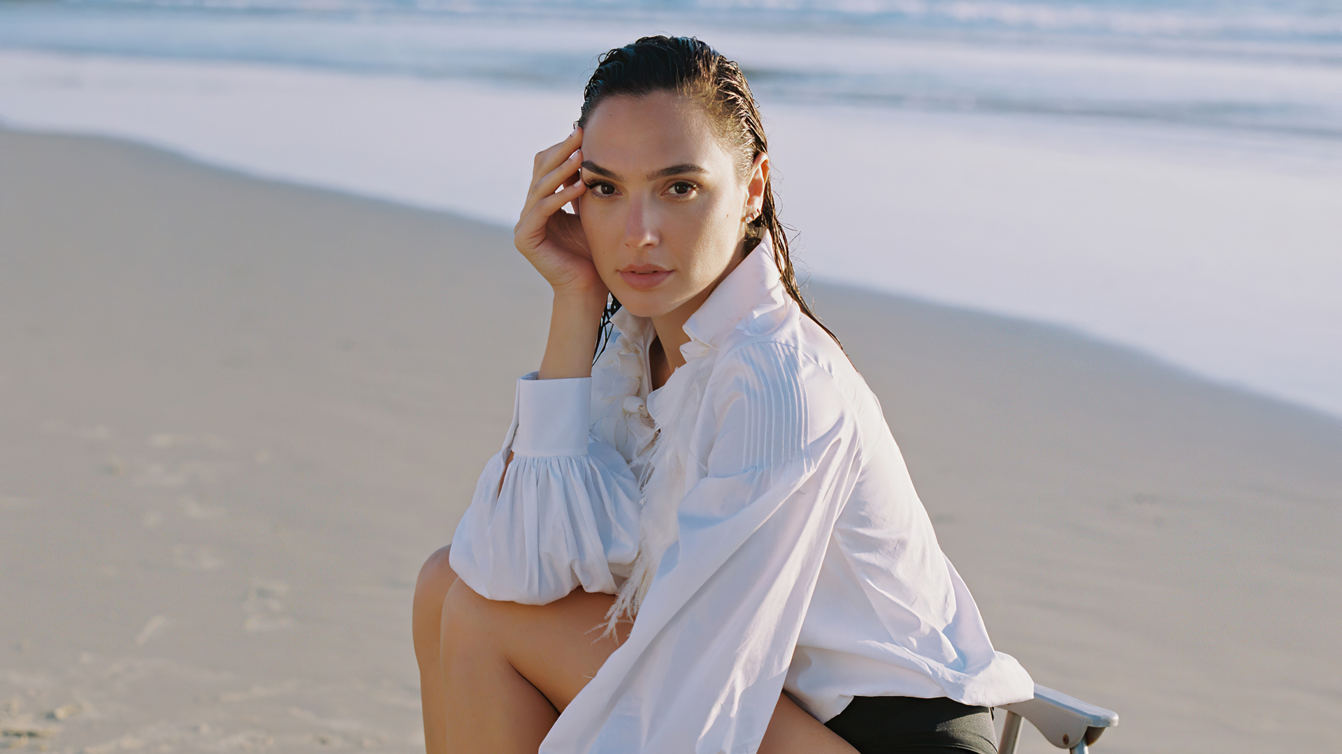Актриса Галь Гадот в белой рубашке сидит на песке