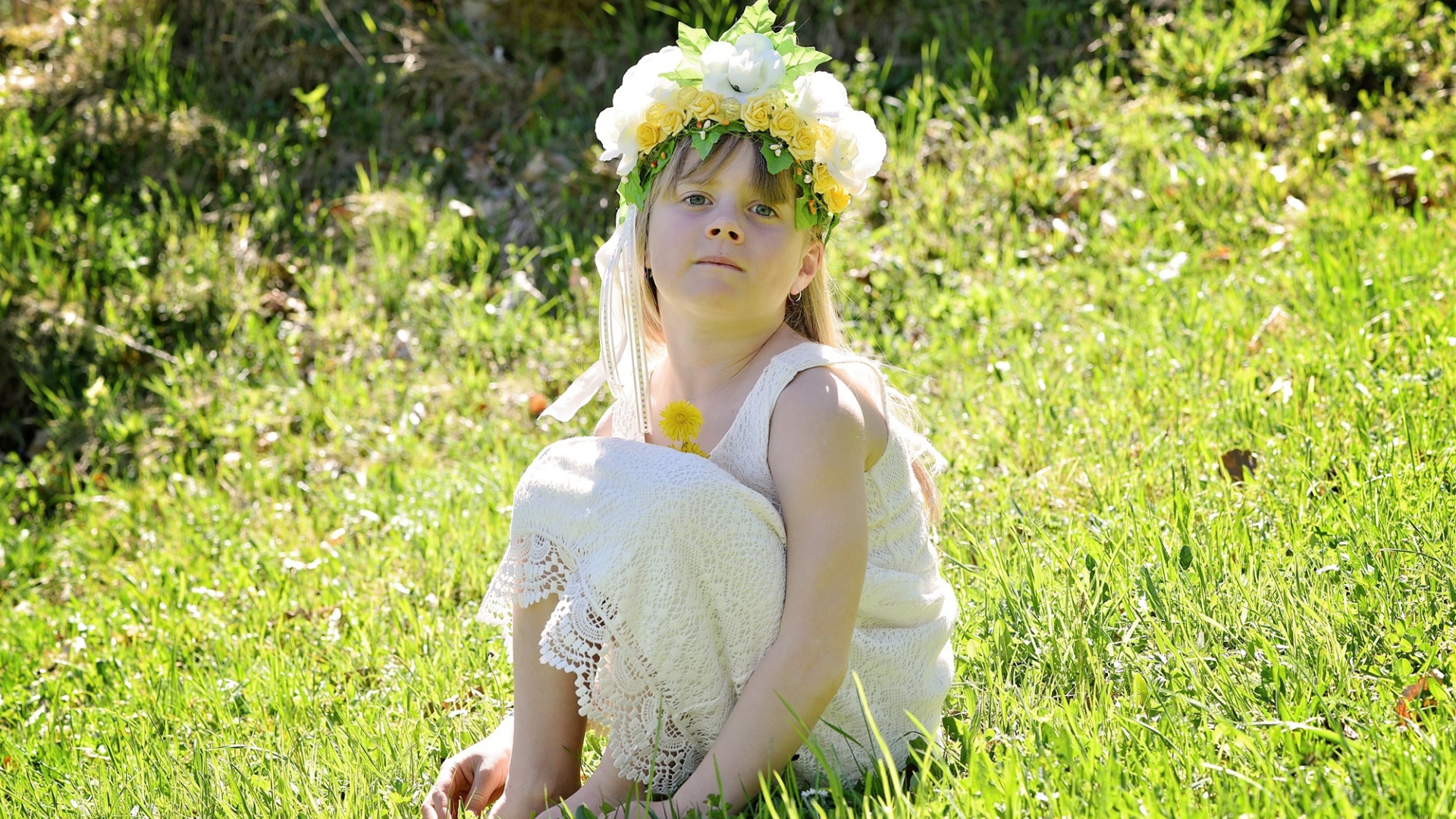 Маленькая девочка с венком на голове сидит на зеленой траве