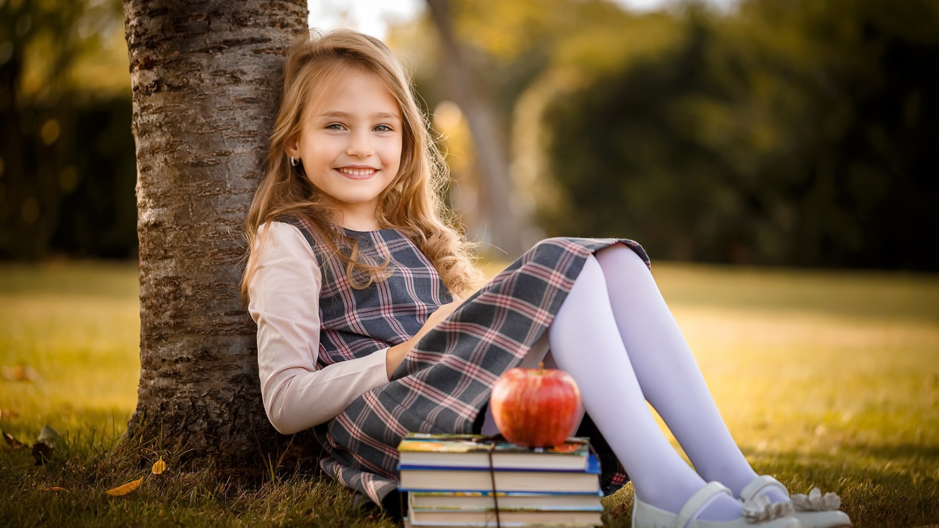 Маленькая девочка с книгами и яблоком сидит под деревом 