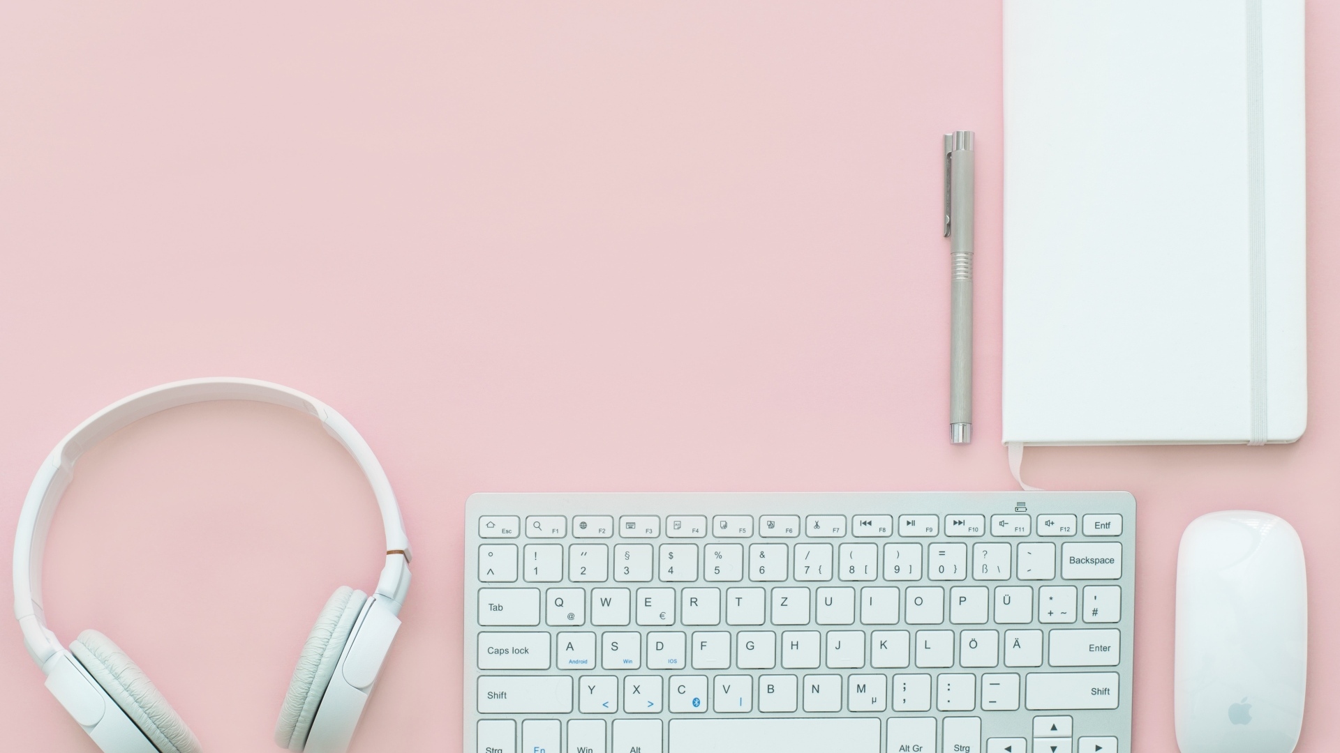 Наушники, клавиатура, блокнот с ручкой и мышка на розовом фоне
