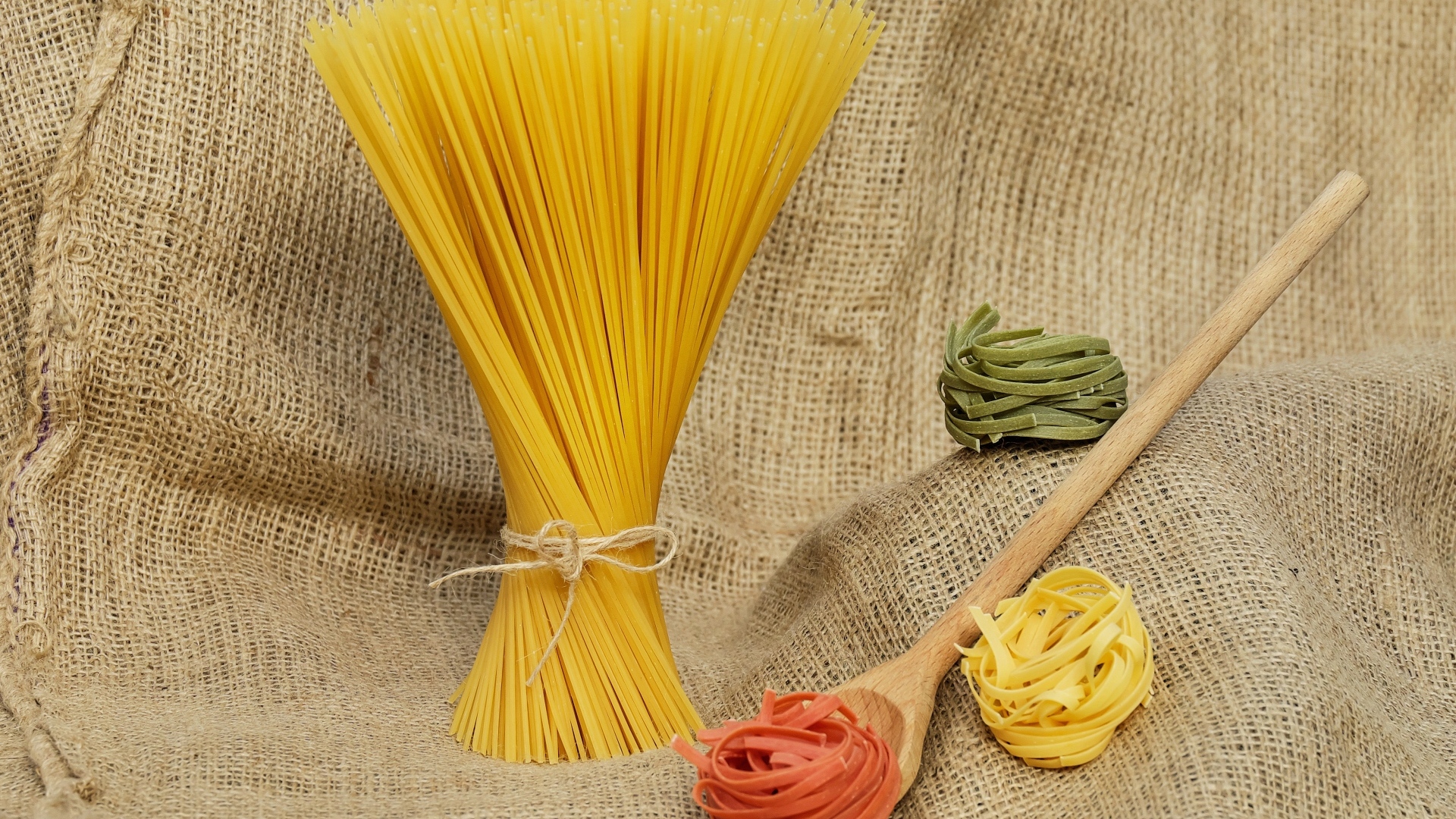 Разноцветные макаронные изделия с деревянной ложкой на мешке