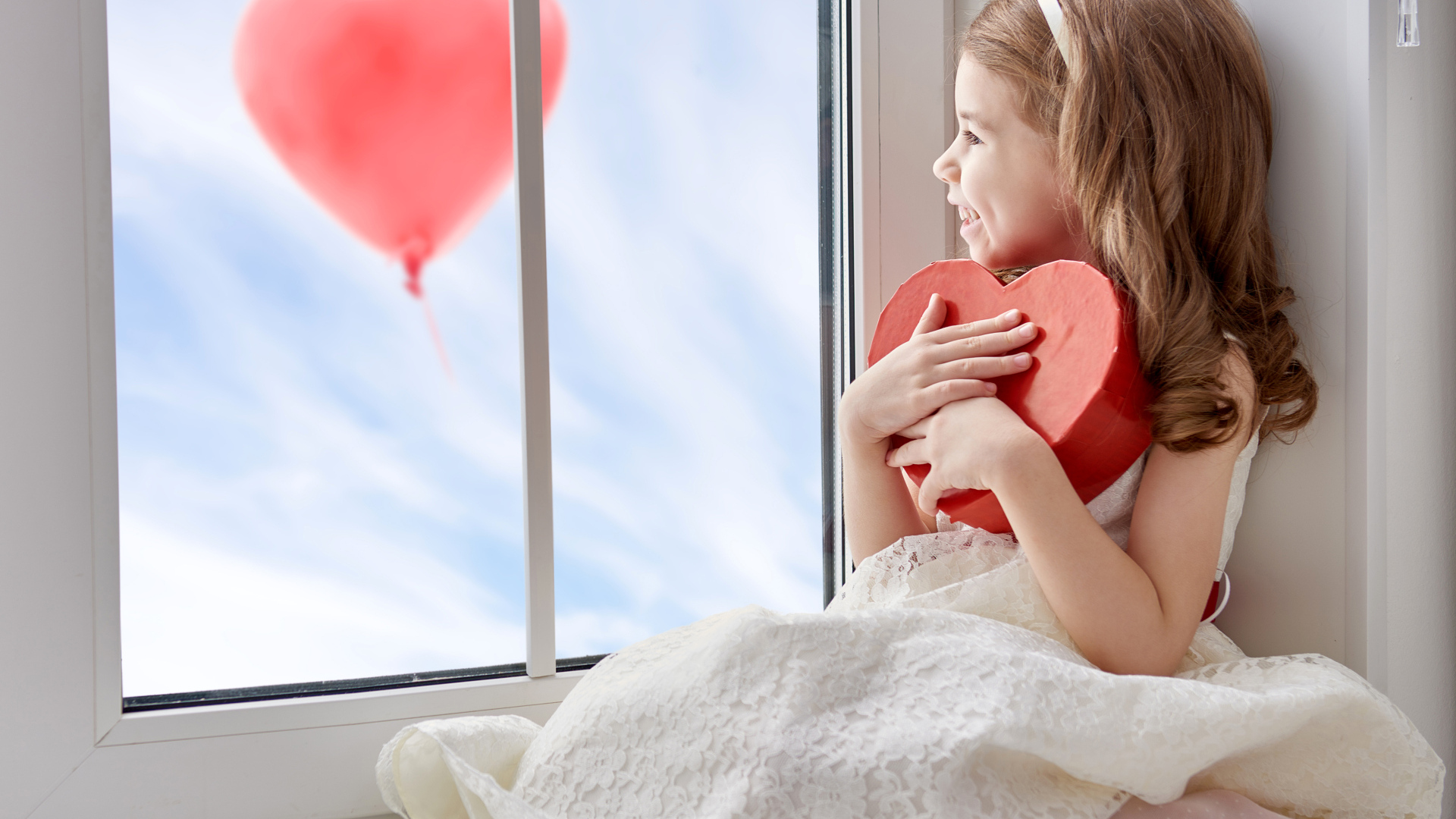 Маленькая девочка с коробкой в форме сердца сидит у окна