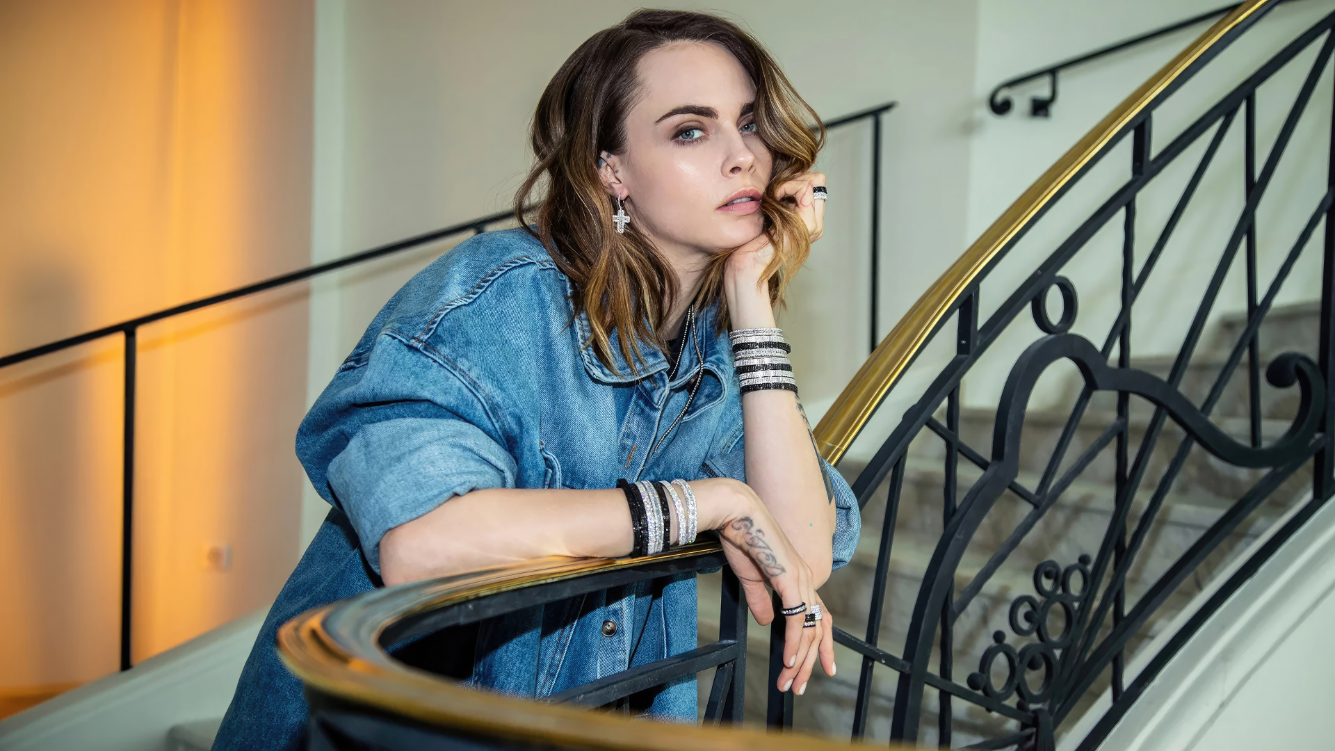 Актриса Кара Делевинь в джинсовой куртке стоит на лестнице