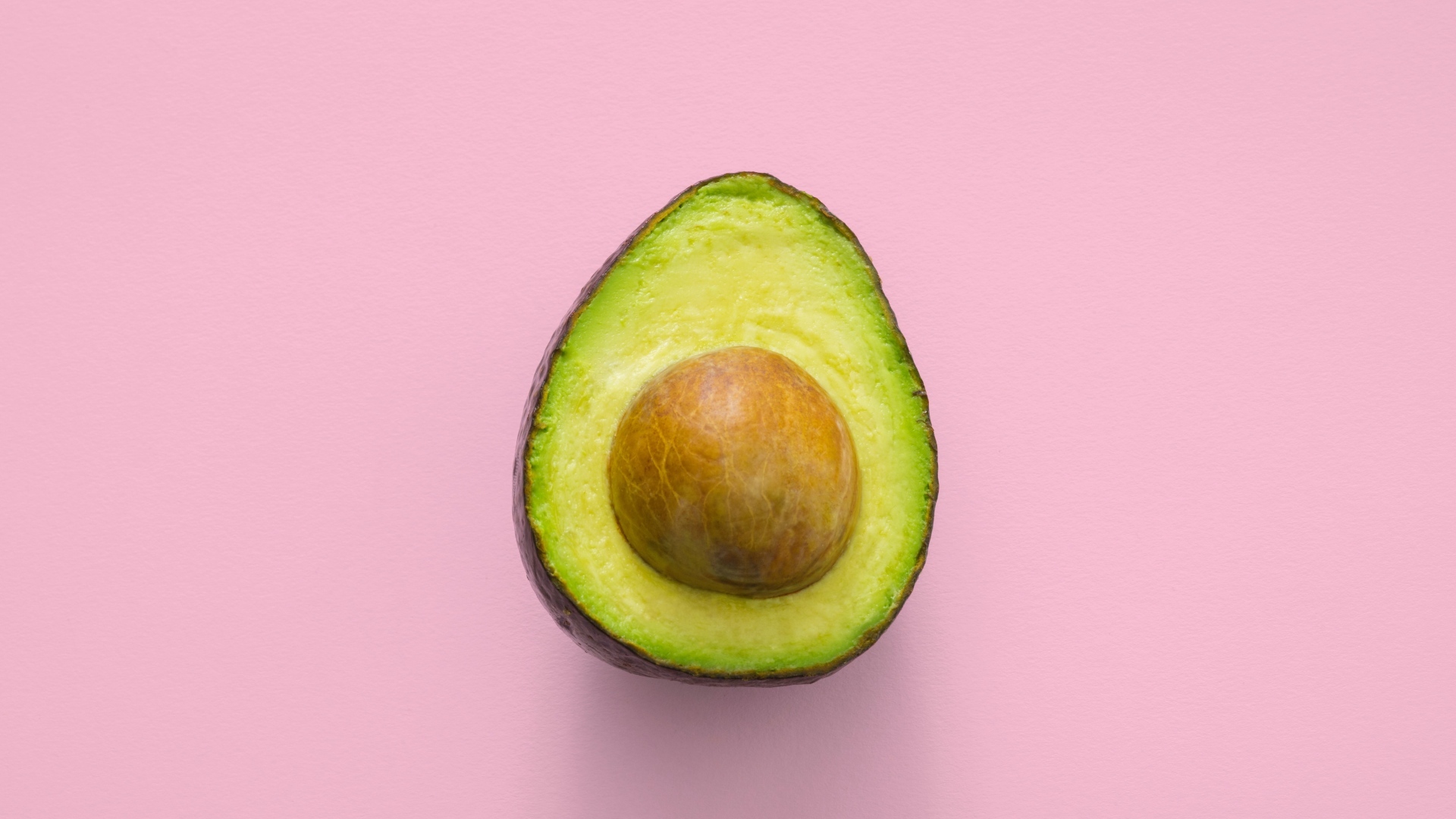 Половина авокадо с большой косточкой на розовом фоне