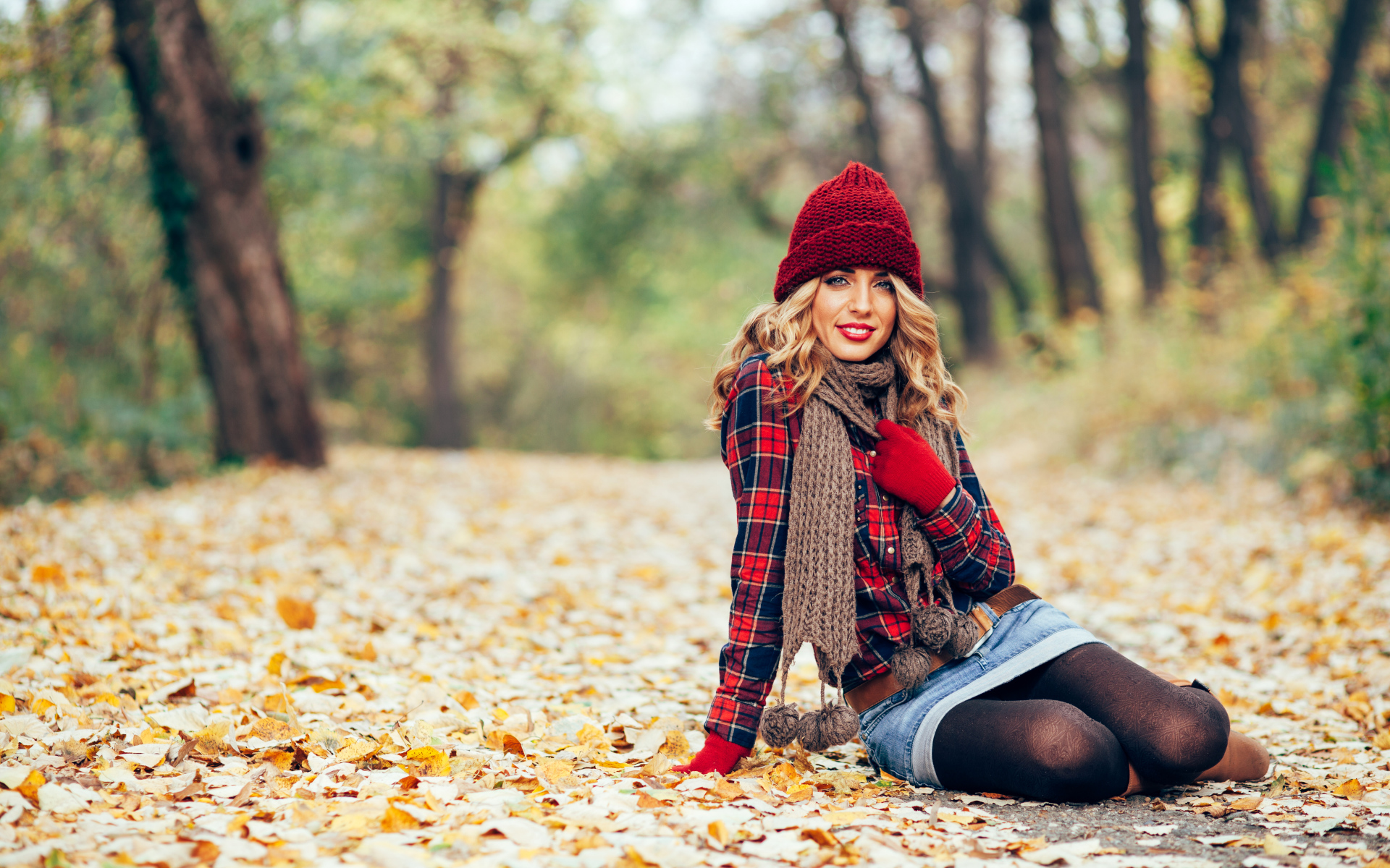 Молодая улыбающаяся девушка в красной шапке сидит на листве осенью