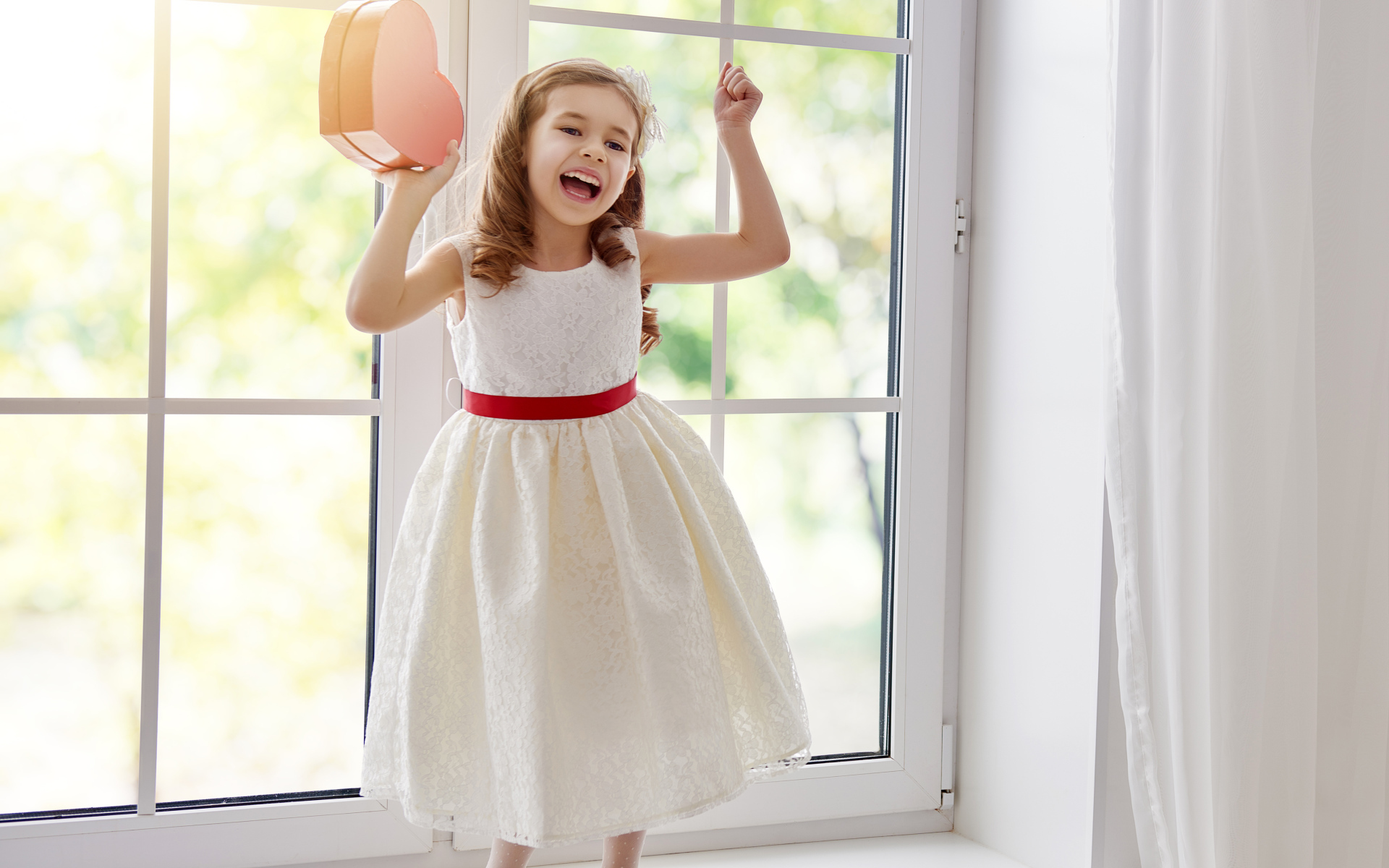 Маленькая девочка с коробкой в форме сердца в руках стоит на подоконнике