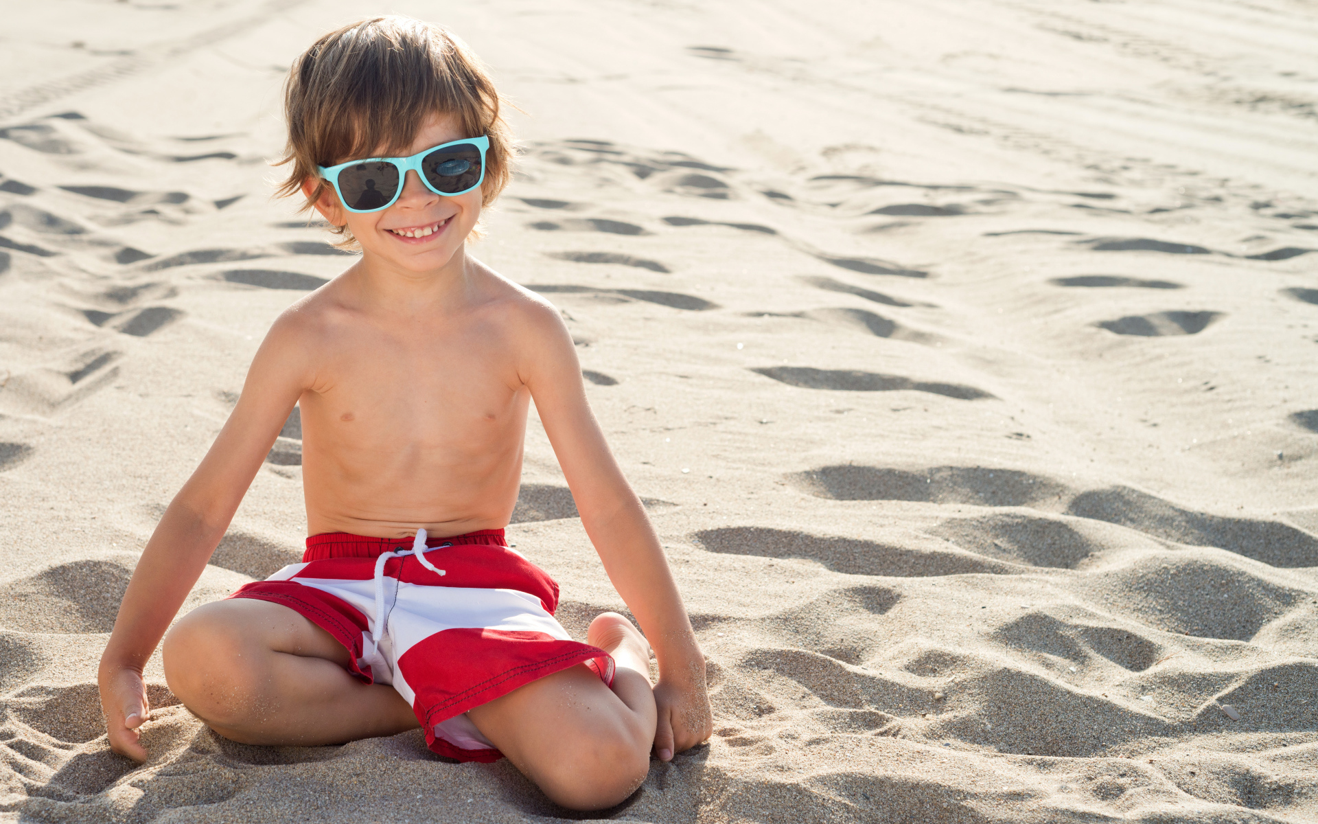 Маленький улыбающийся мальчик в очках сидит на песке