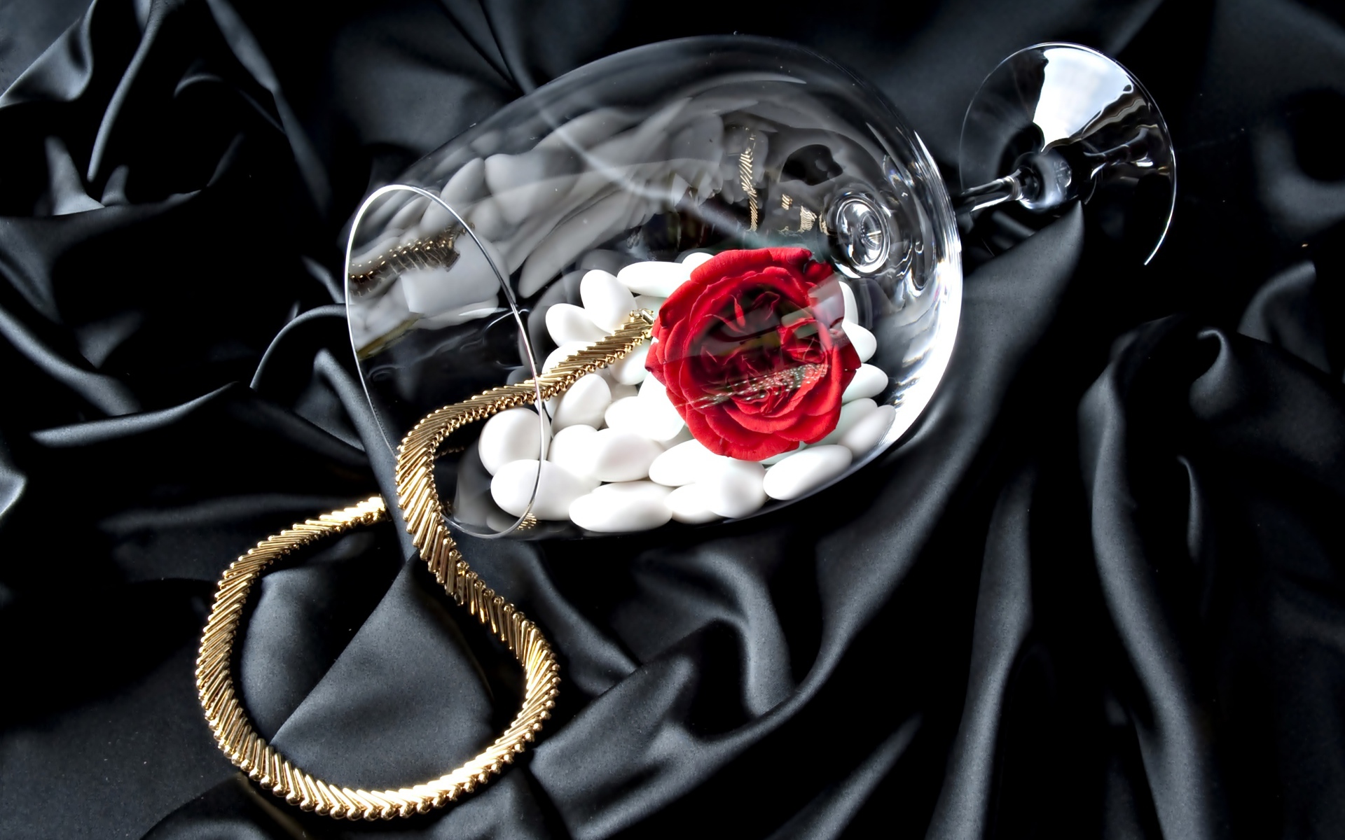 Бокал с золотой цепочкой, красной розой и белыми камнями на черном покрывале