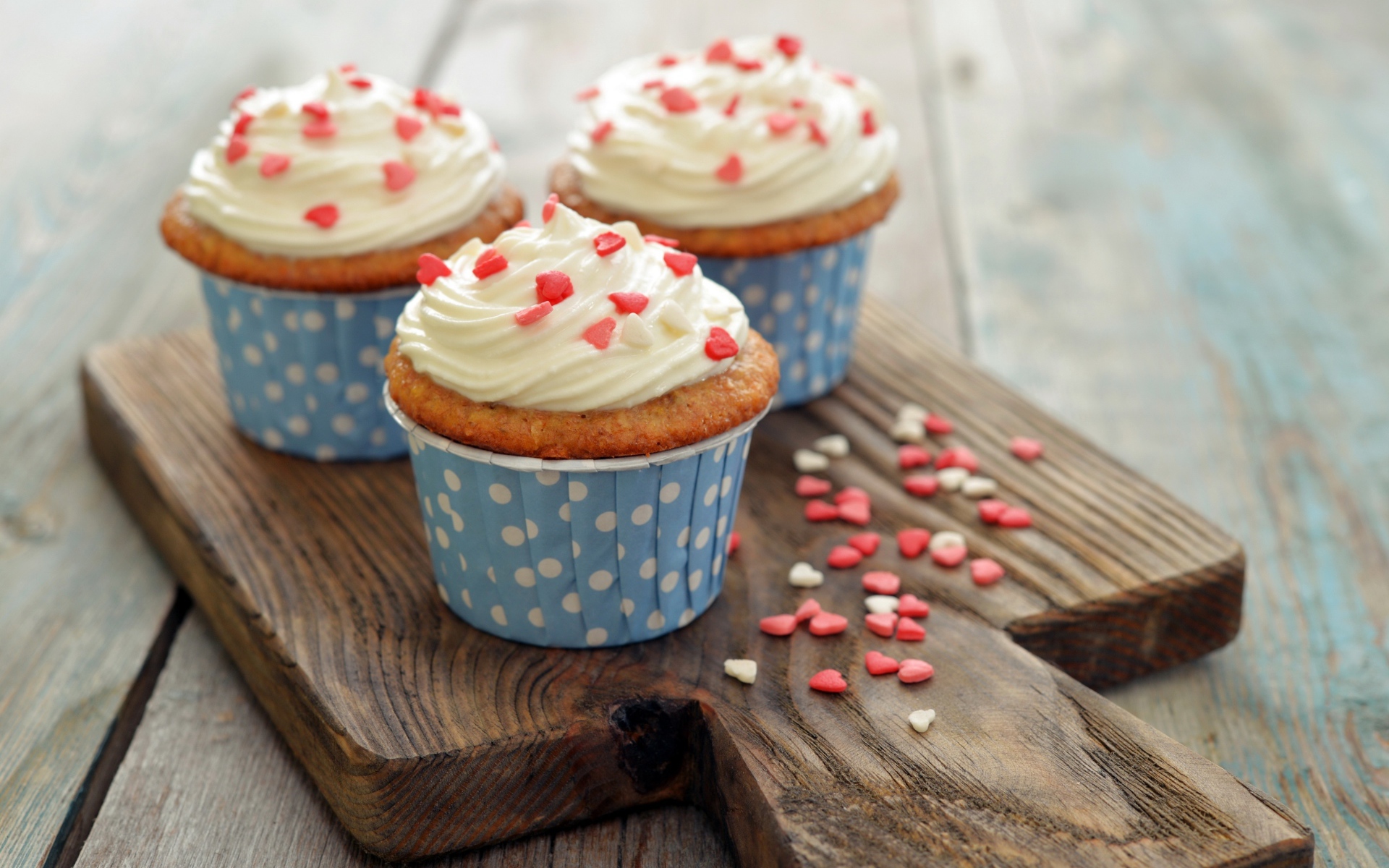 Три кекса со сливками украшены сердечками стоят на разделочной доске