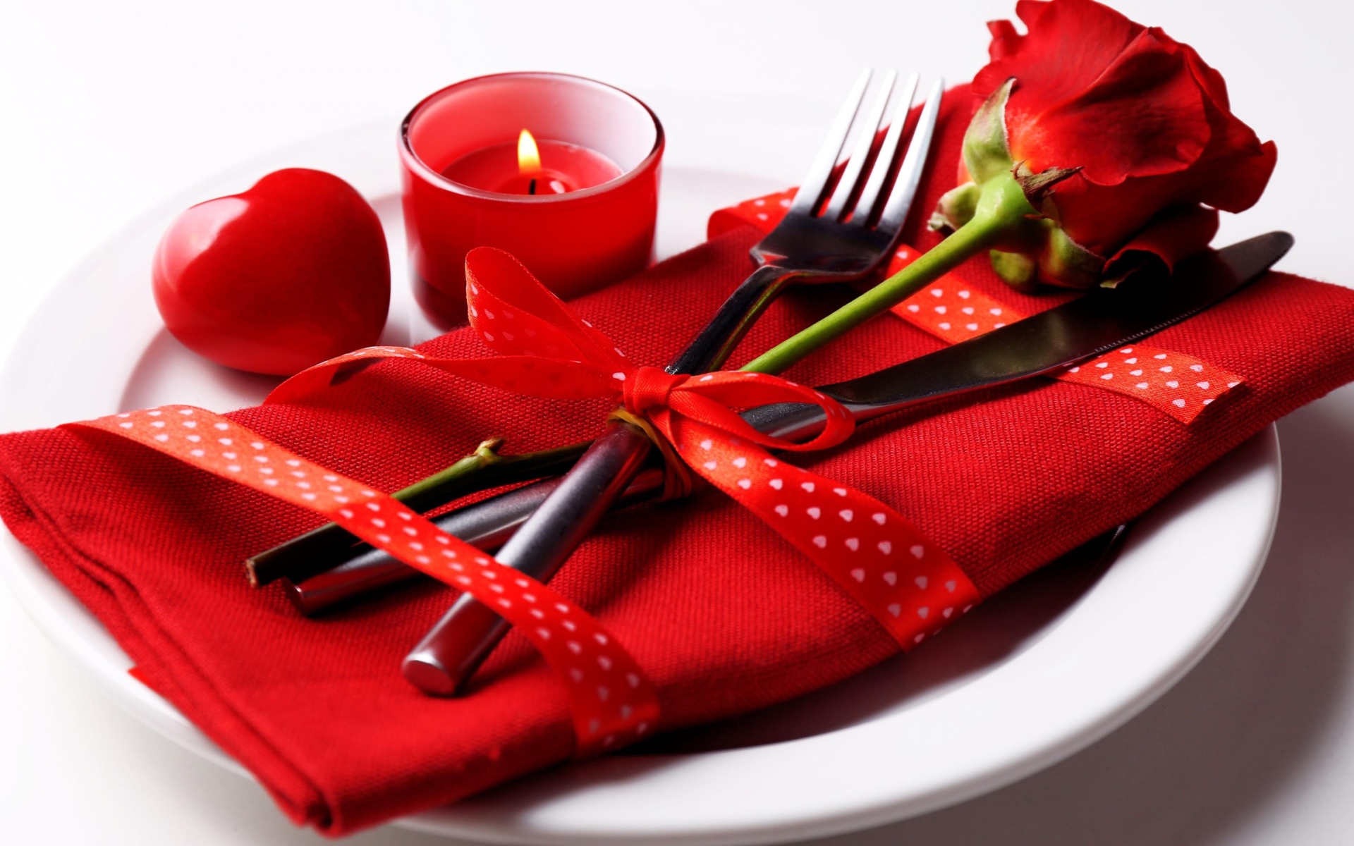 Роза, столовые приборы и зажженная свеча на белой тарелке 