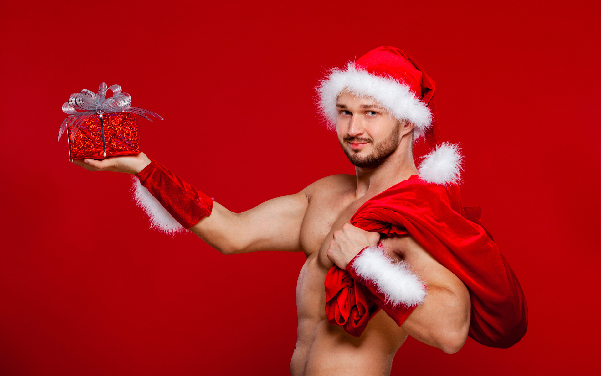 Мужчина в новогоднем костюме Санта Клауса с мешком и подарком в руке на красном фоне