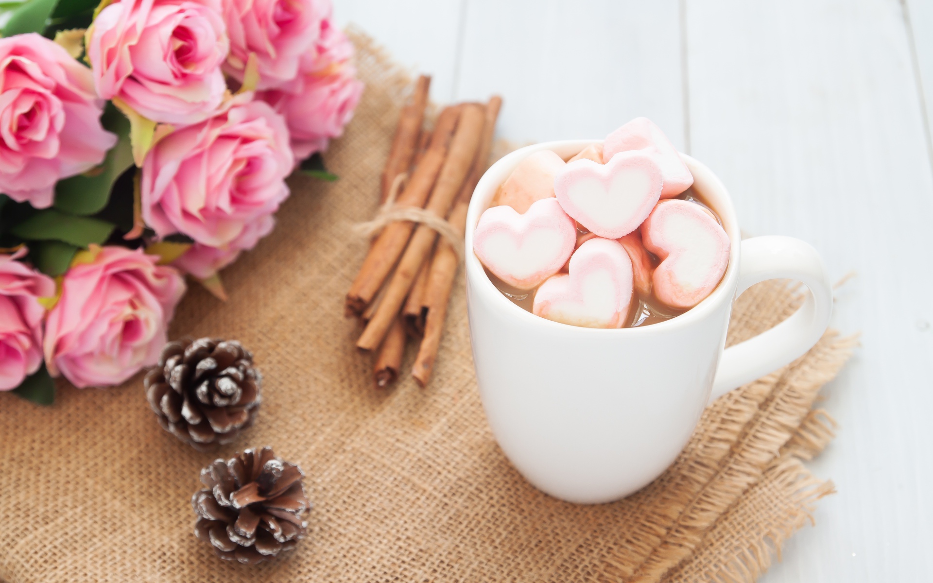 Чашка какао с маршмеллоу на столе с корицей, шишками и букетом розовых роз