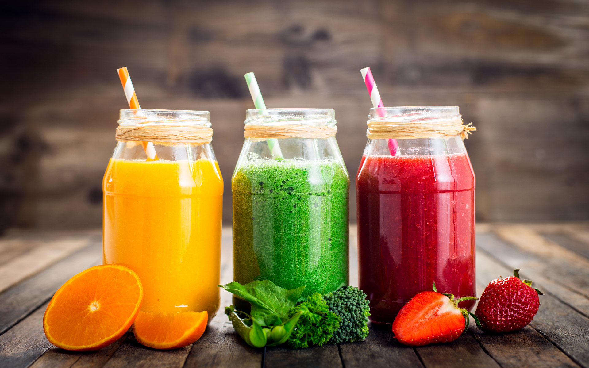 Три банки смузи с трубочкой на столе с апельсином, брокколи и клубникой