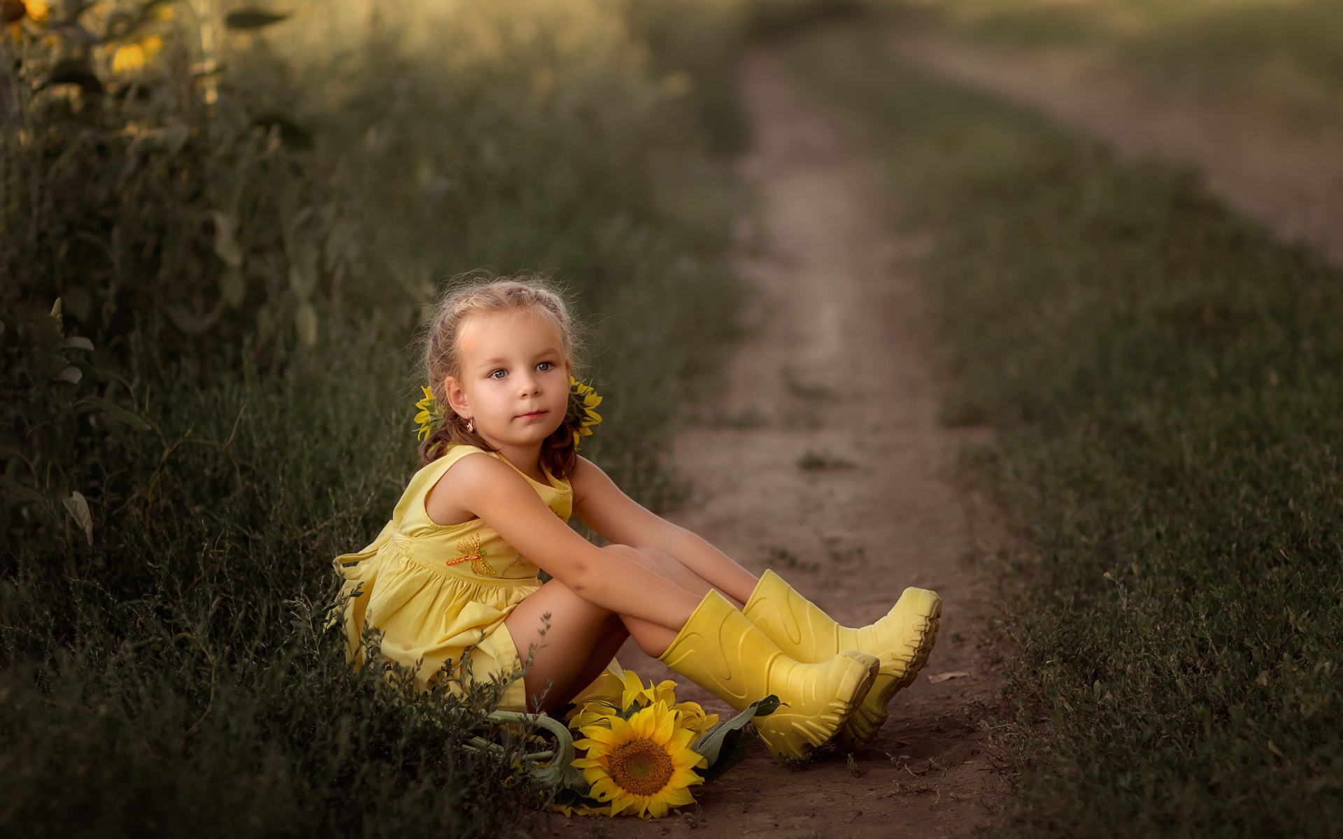 Маленькая девочка в желтом платье сидит на земле с подсолнухами