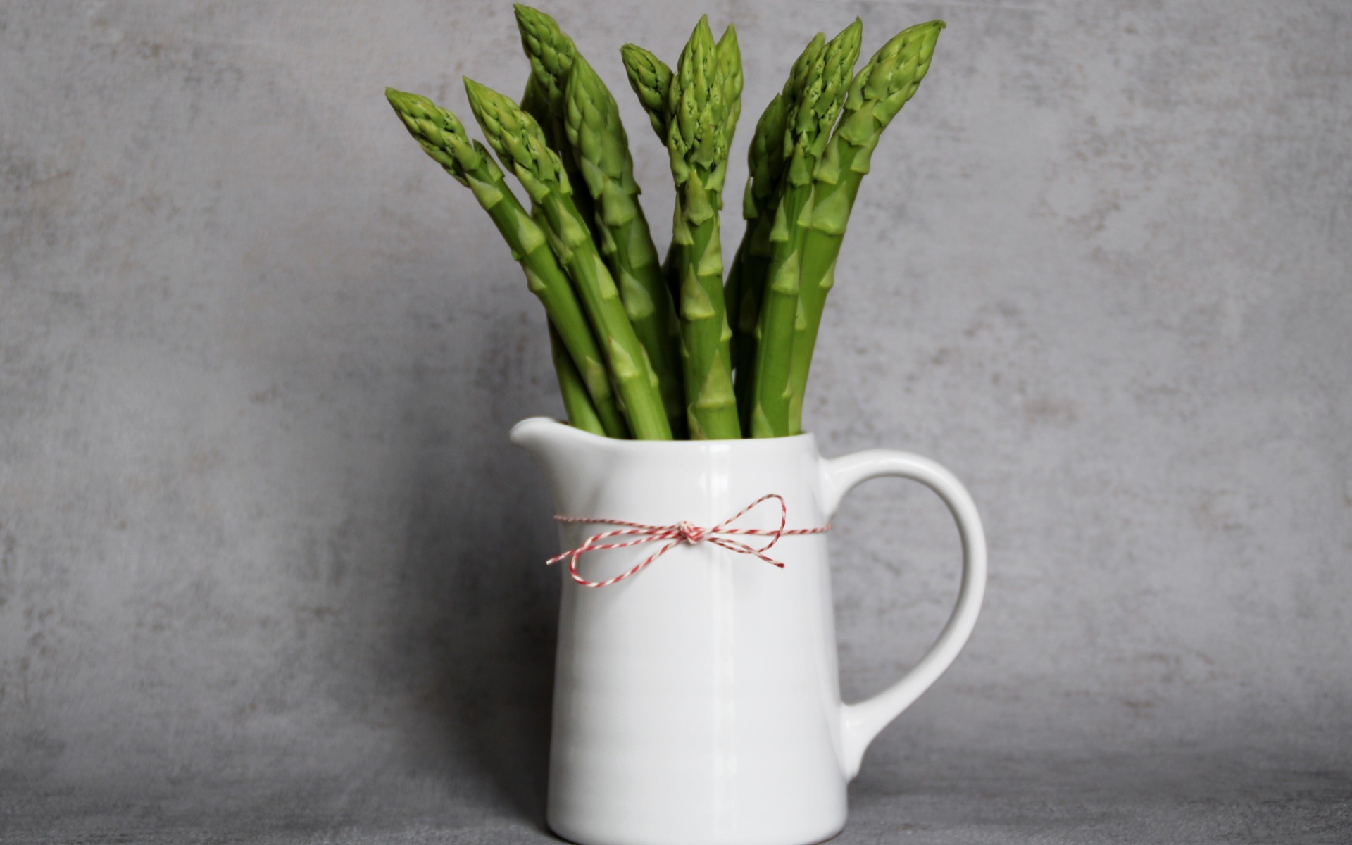 Зеленая спаржа в белой вазе на фоне серой стены