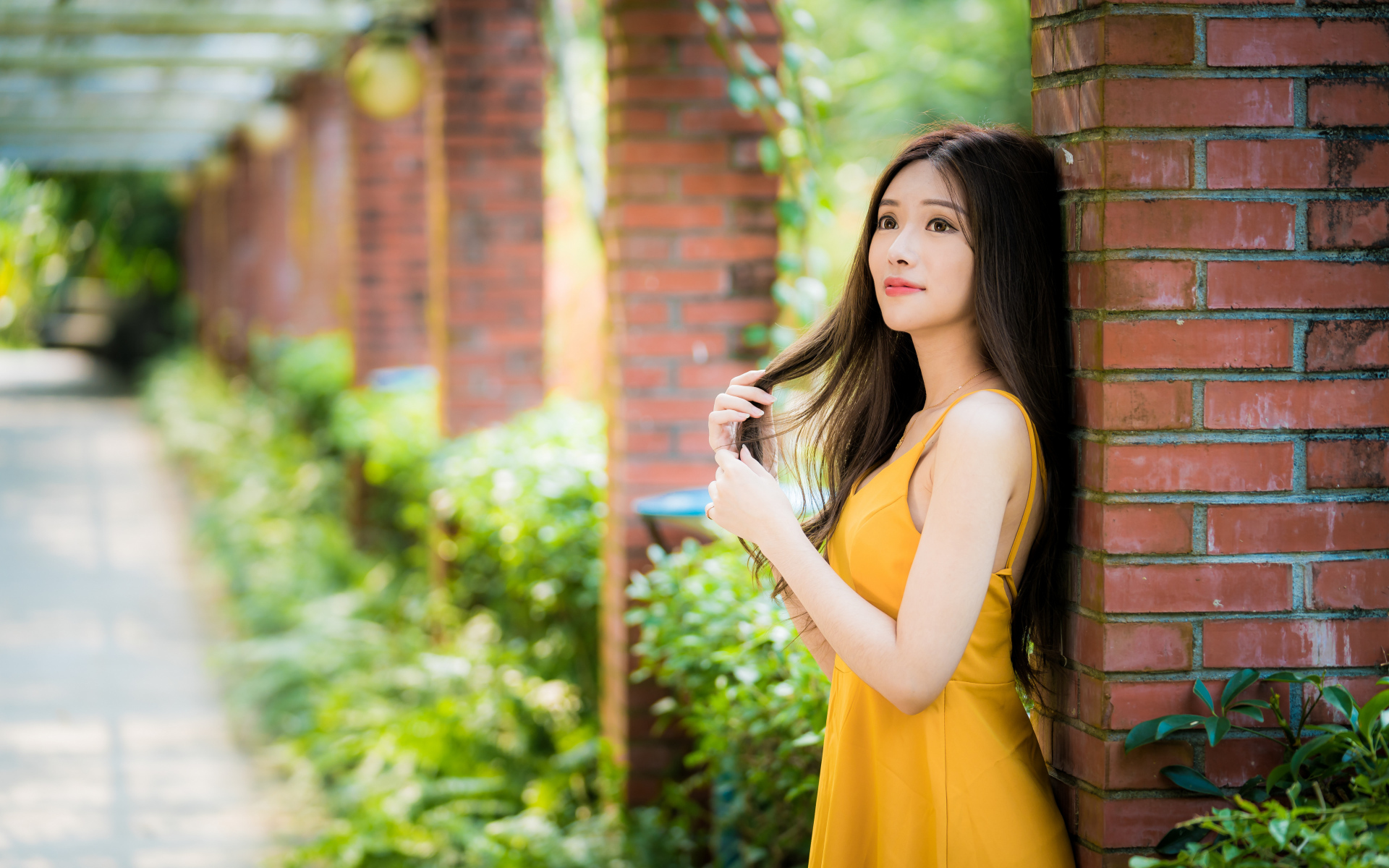 Красивая азиатка в желтом платье стоит у кирпичной колонны 
