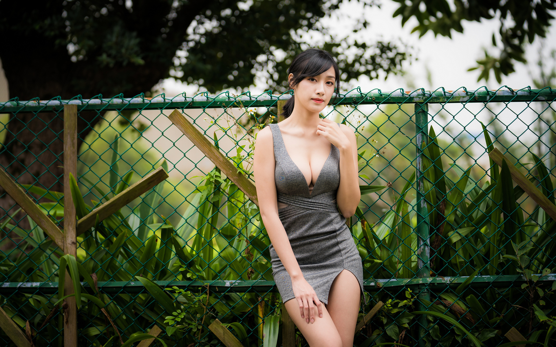 Красивая азиатка в сером платье стоит у зеленой сетки 
