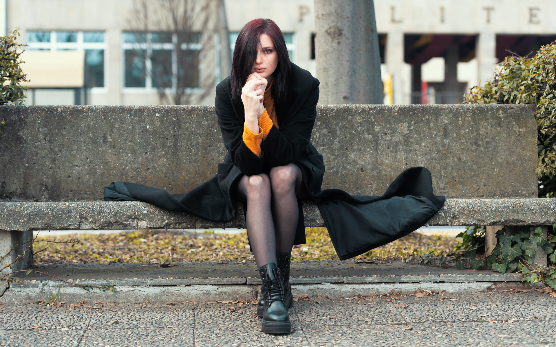 Девушка в черном пальто сидит на бетонной лавке 