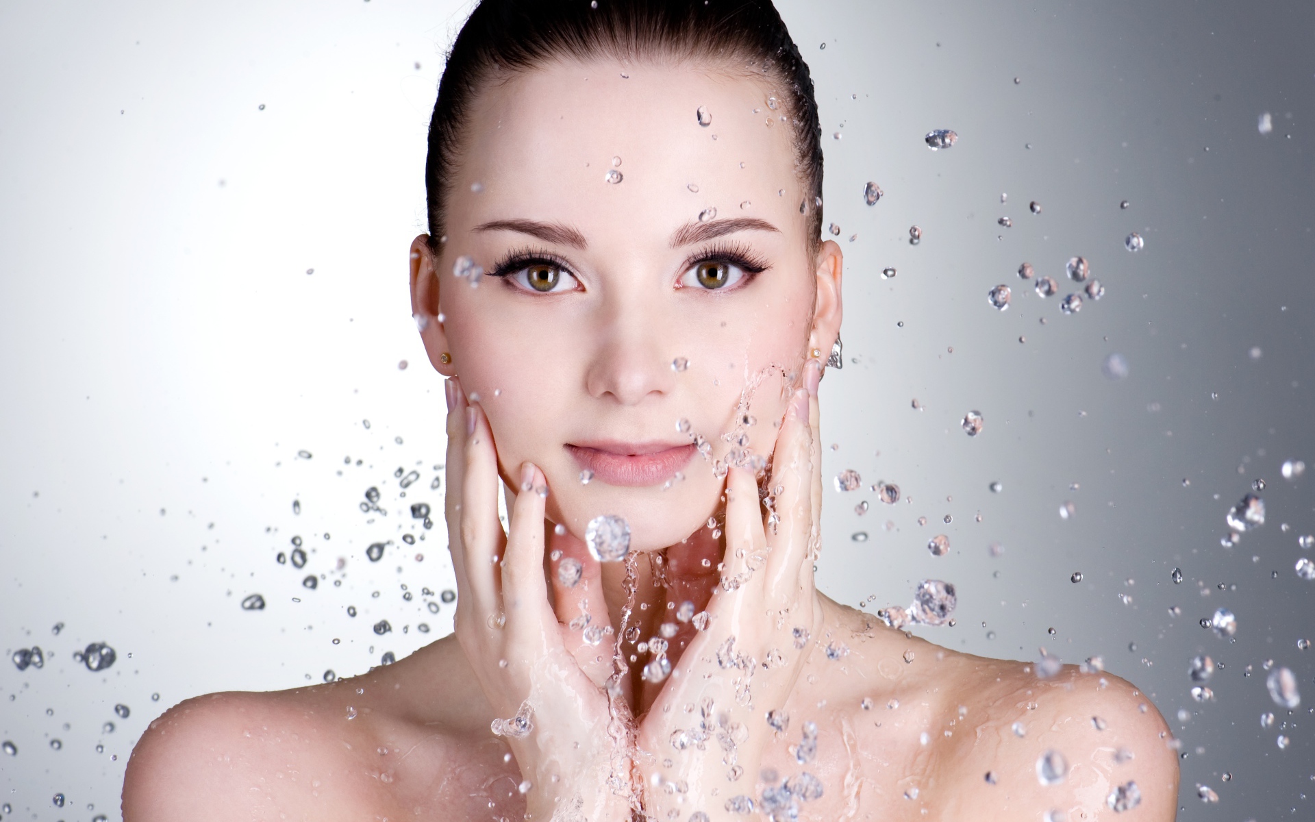 Брызги воды летят на красивое лицо молодой девушки на сером фоне