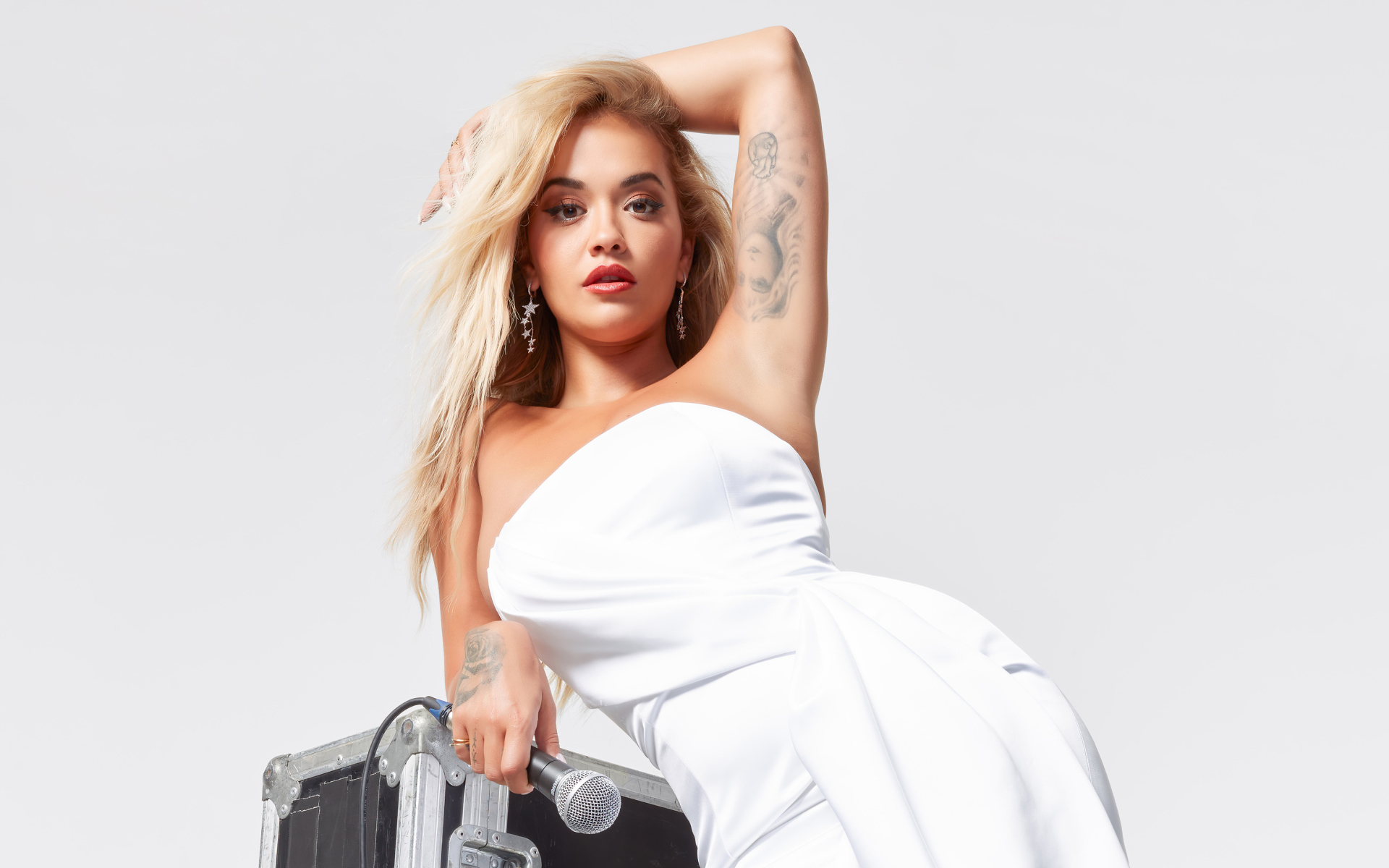 Певица Рита Ора в белом платье с микрофоном на сером фоне 